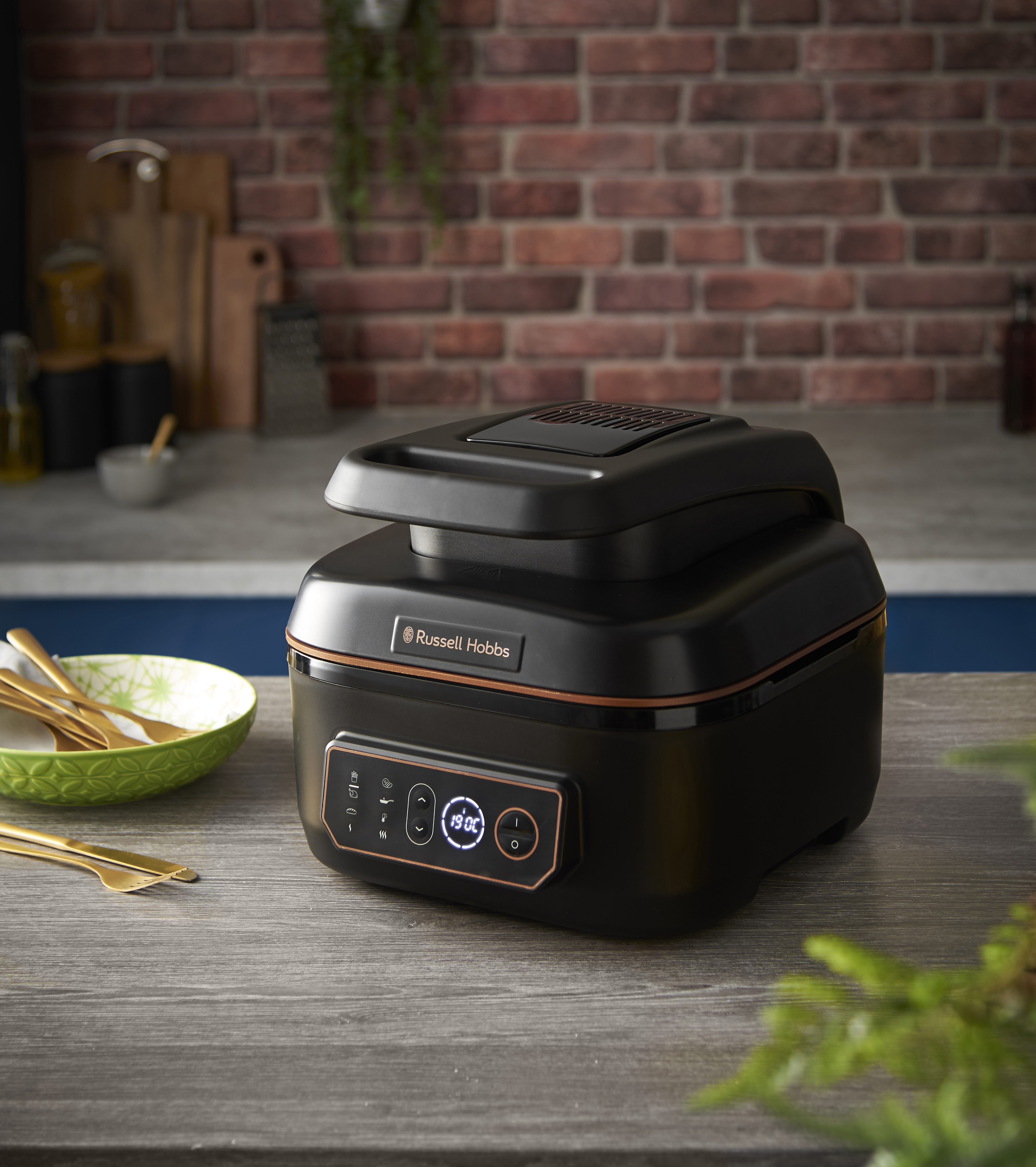 Fekete Russell Hobbs légkeveréses sütő fa felületen, téglával a háttérben. Digitális kijelzővel.