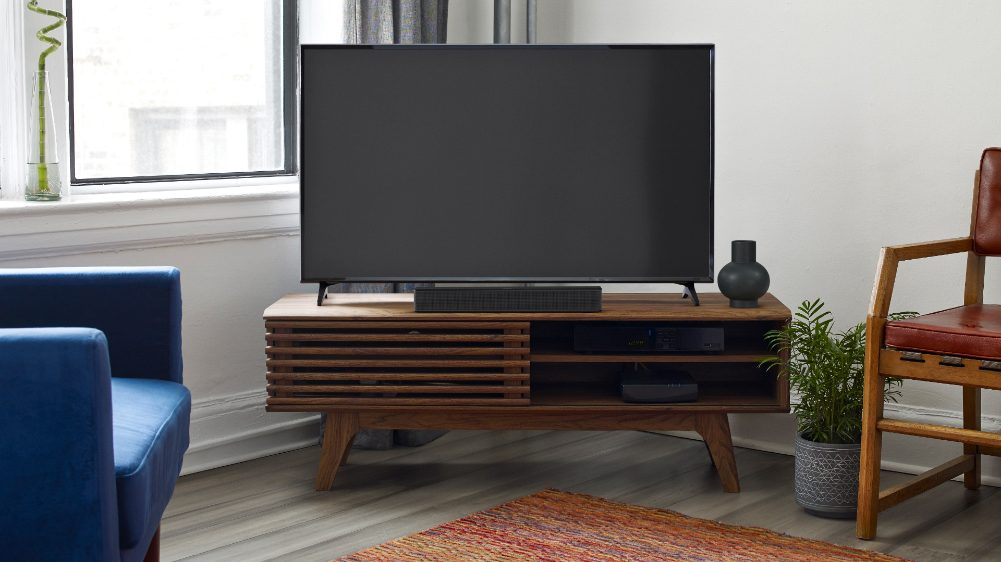Een tv staat op een houten standaard met een soundbar. Een blauwe bank en stoel zijn zichtbaar.