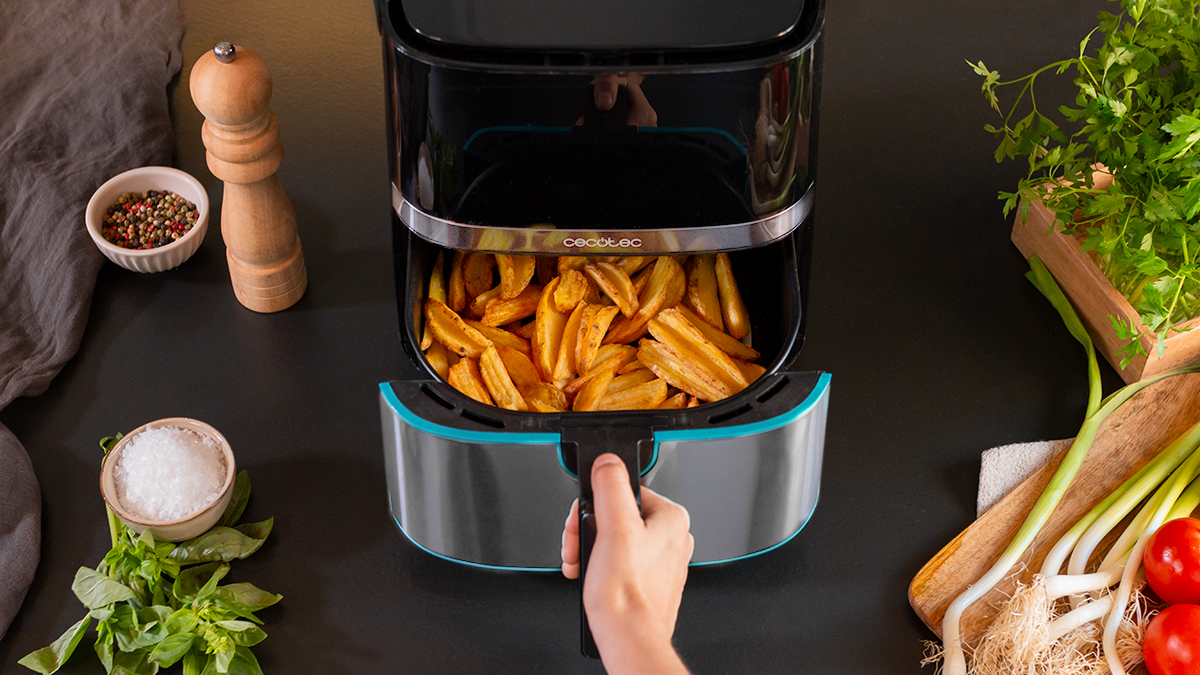 Freidora de aire con patatas fritas dentro y una persona sosteniendo el mango. Encimera negra, ingredientes.