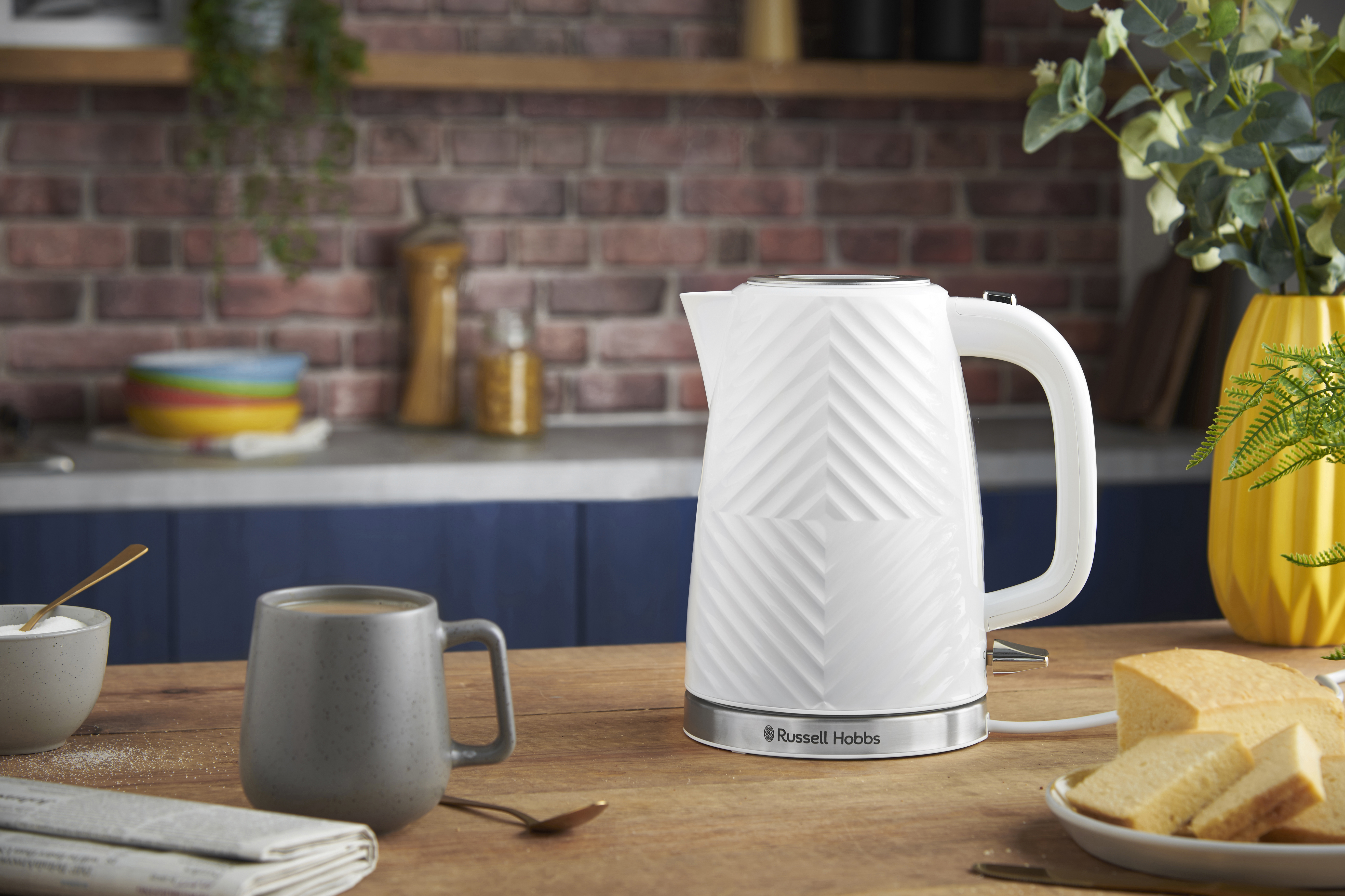 Egy fehér Russell Hobbs vízforraló egy faasztalon kávéval és kenyérrel.