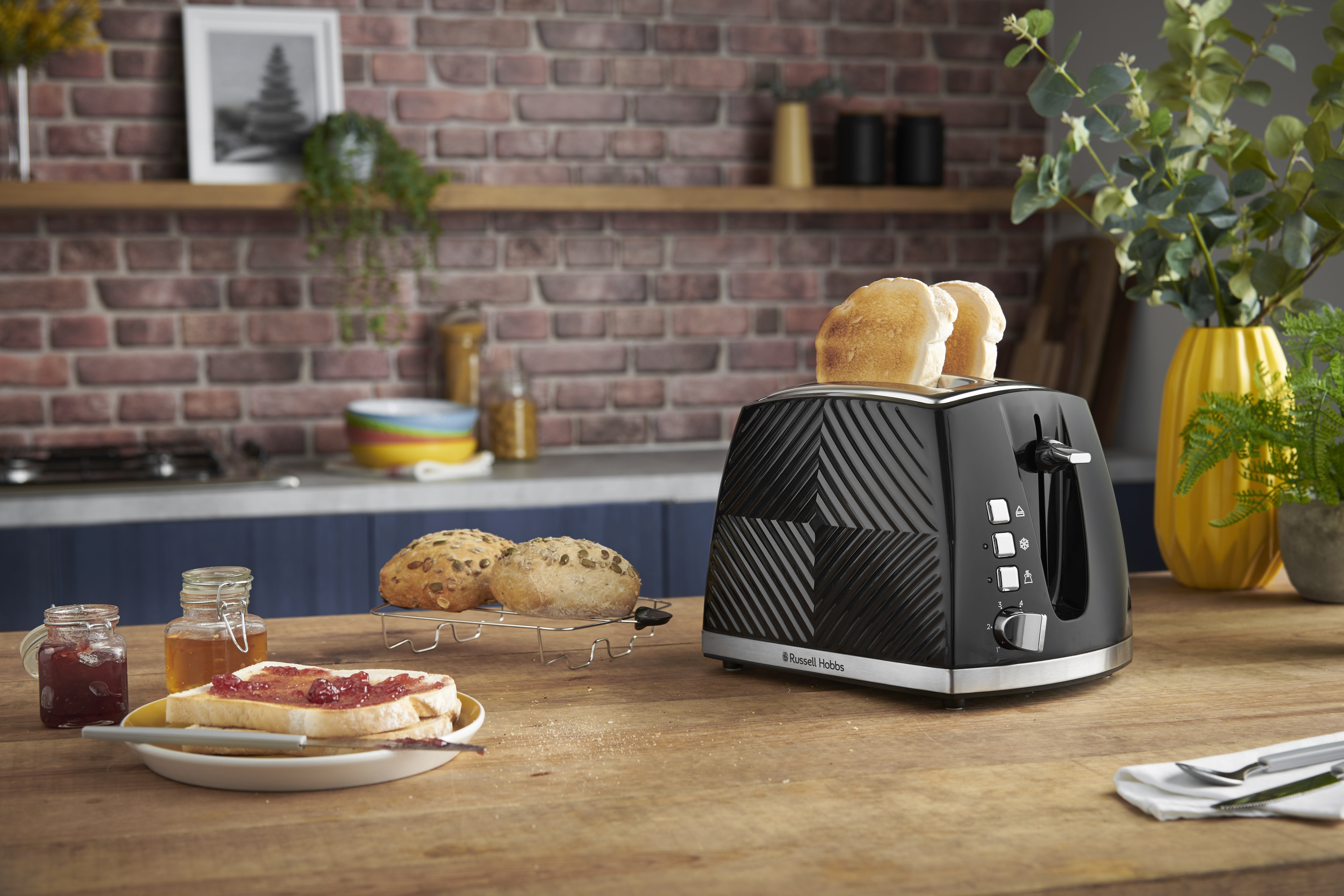 Fekete Russell Hobbs kenyérpirító, két pirítósszelettel. Valamint lekváros pirítós és zsemlék egy faasztalon.