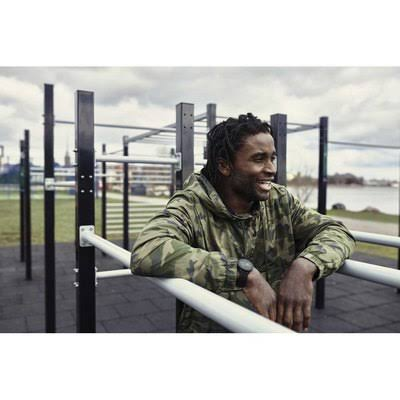 Un hombre con chaqueta camuflada apoyado en una estructura de barras metálicas, sonriendo. Gimnasio al aire libre con un lago.