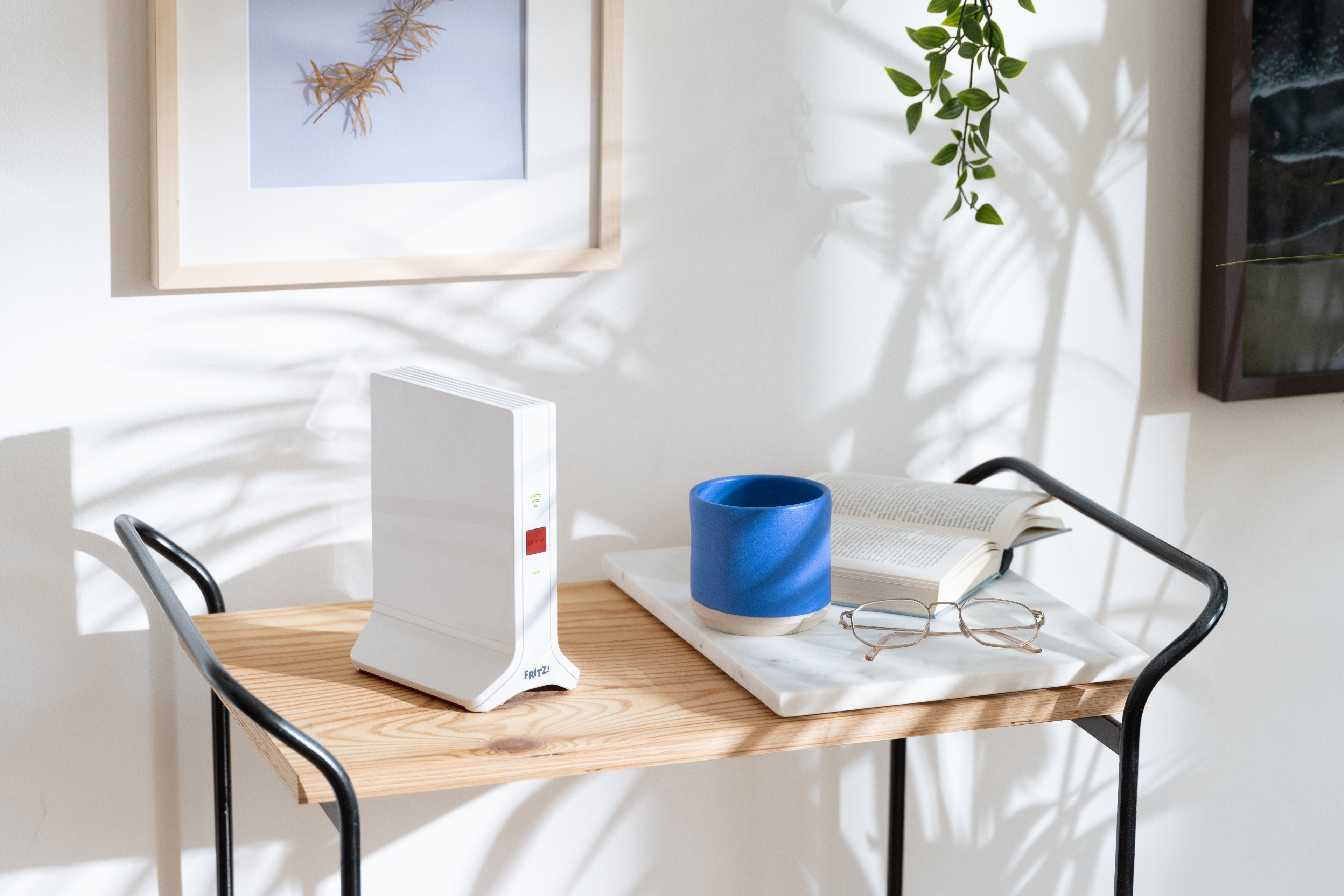Tisch mit Router, blauer Tasse, Buch und Brille. Bilderrahmen und Pflanze im Hintergrund.