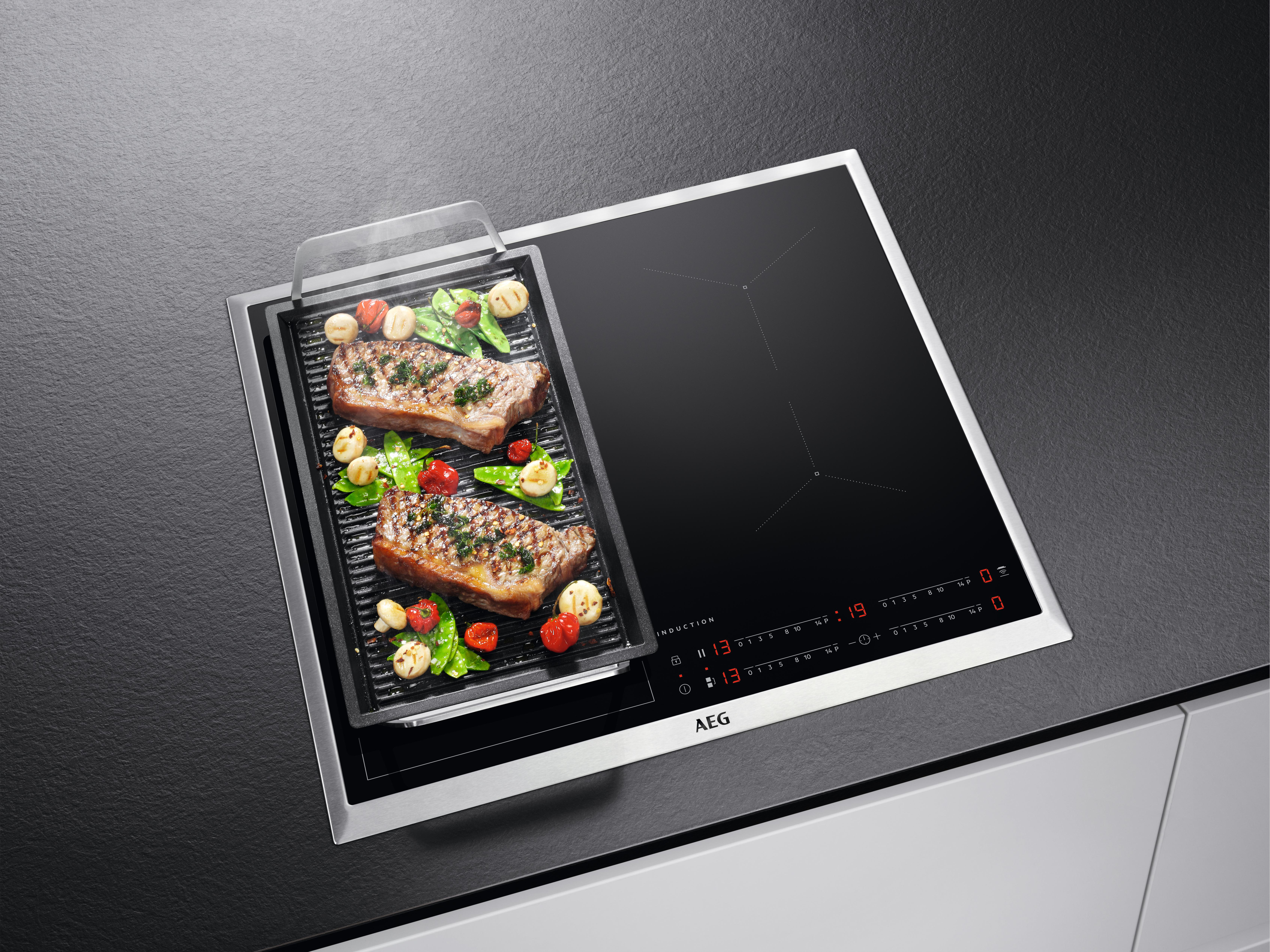 Schwarzes Kochfeld mit gegrillten Steaks und Gemüse. Das Kochfeld hat digitale Bedienelemente. Der Hintergrund ist dunkelgrau.