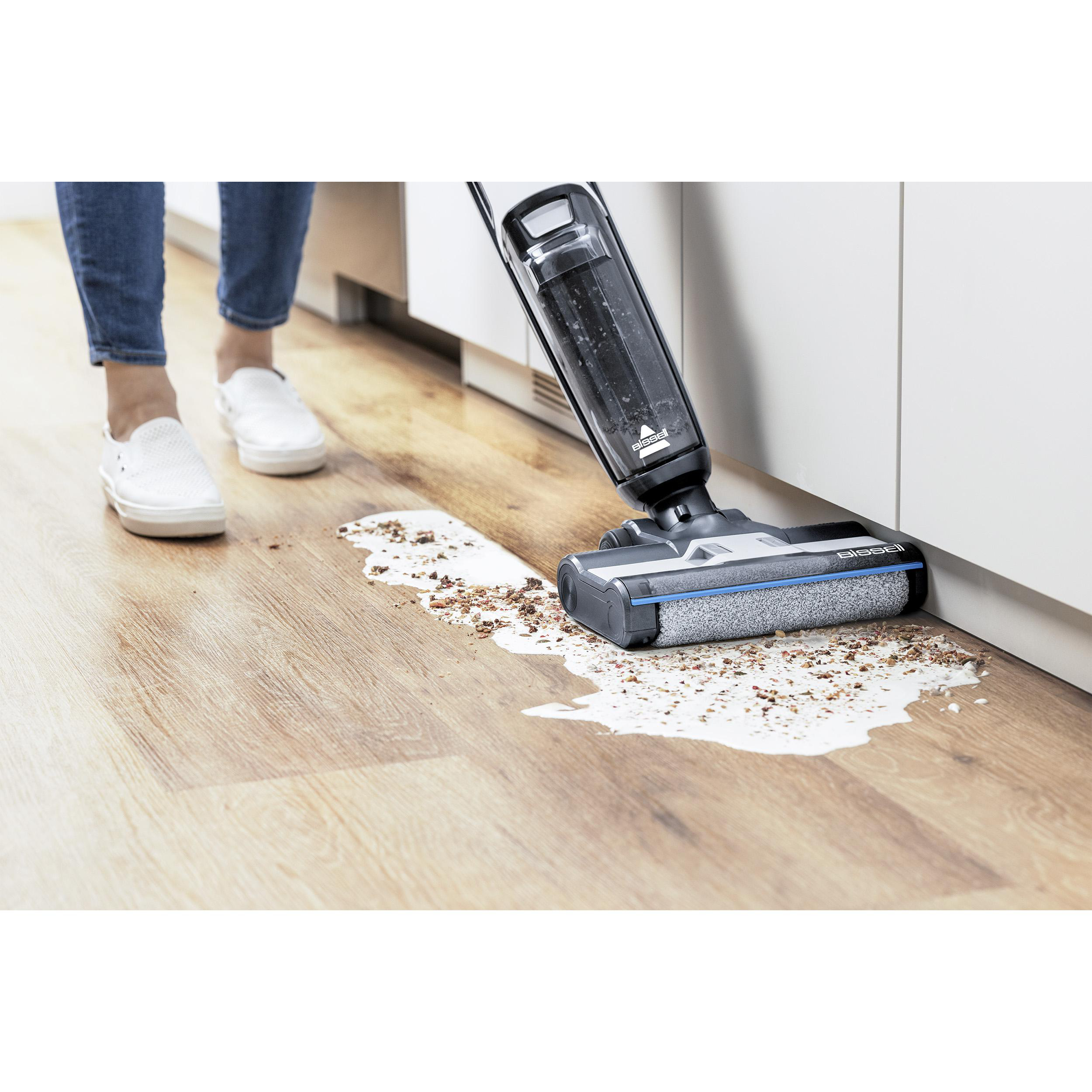 Person reinigt einen verschütteten Fleck auf einem Holzboden mit einem Staubsauger. Weiße Schuhe und Schränke sind sichtbar.