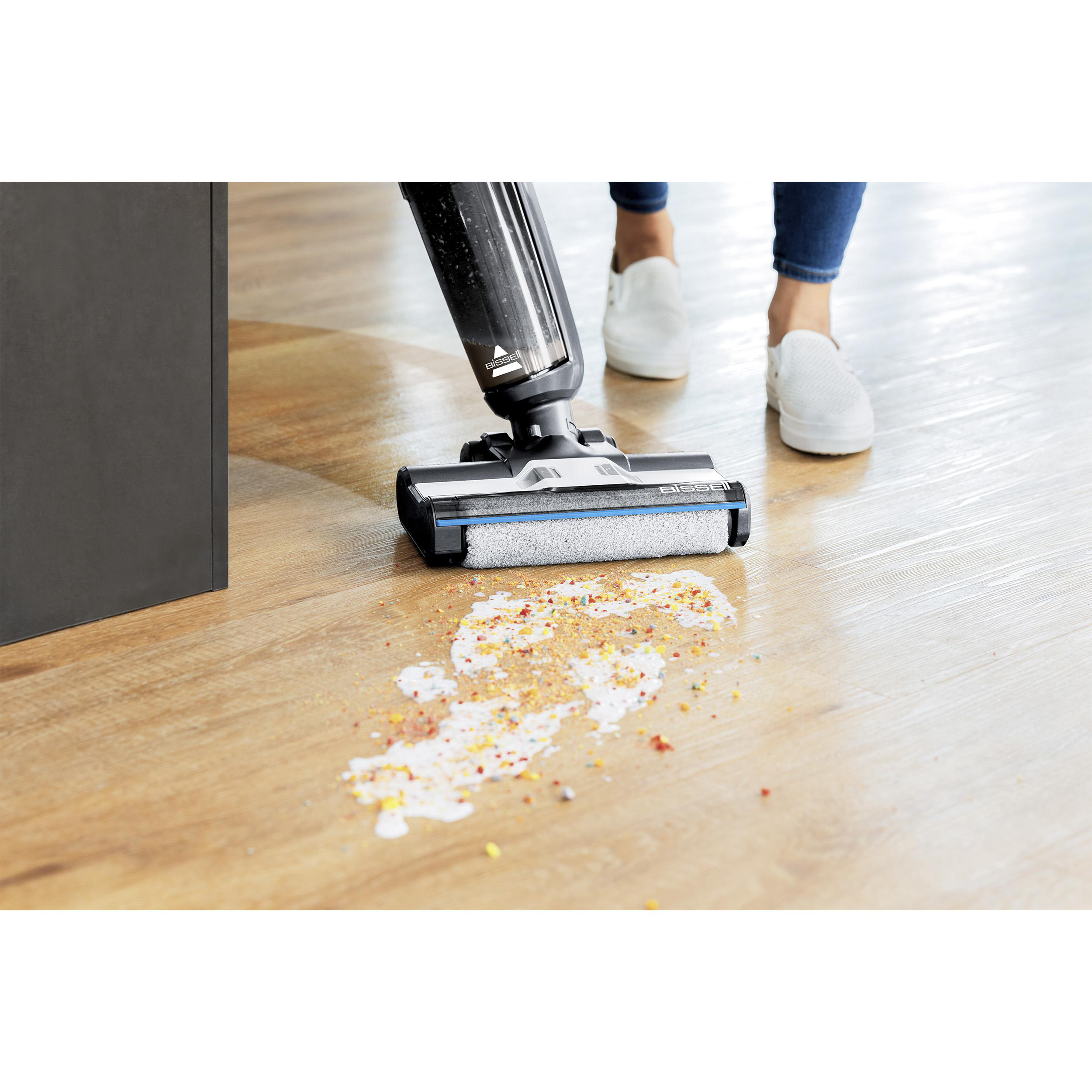 Eine Person reinigt einen verschütteten Flüssigkeitsfleck mit einem Staubsauger. Der Fleck ist eine Mischung aus Flüssigkeiten und Streuseln. Der Boden ist aus Holz.
