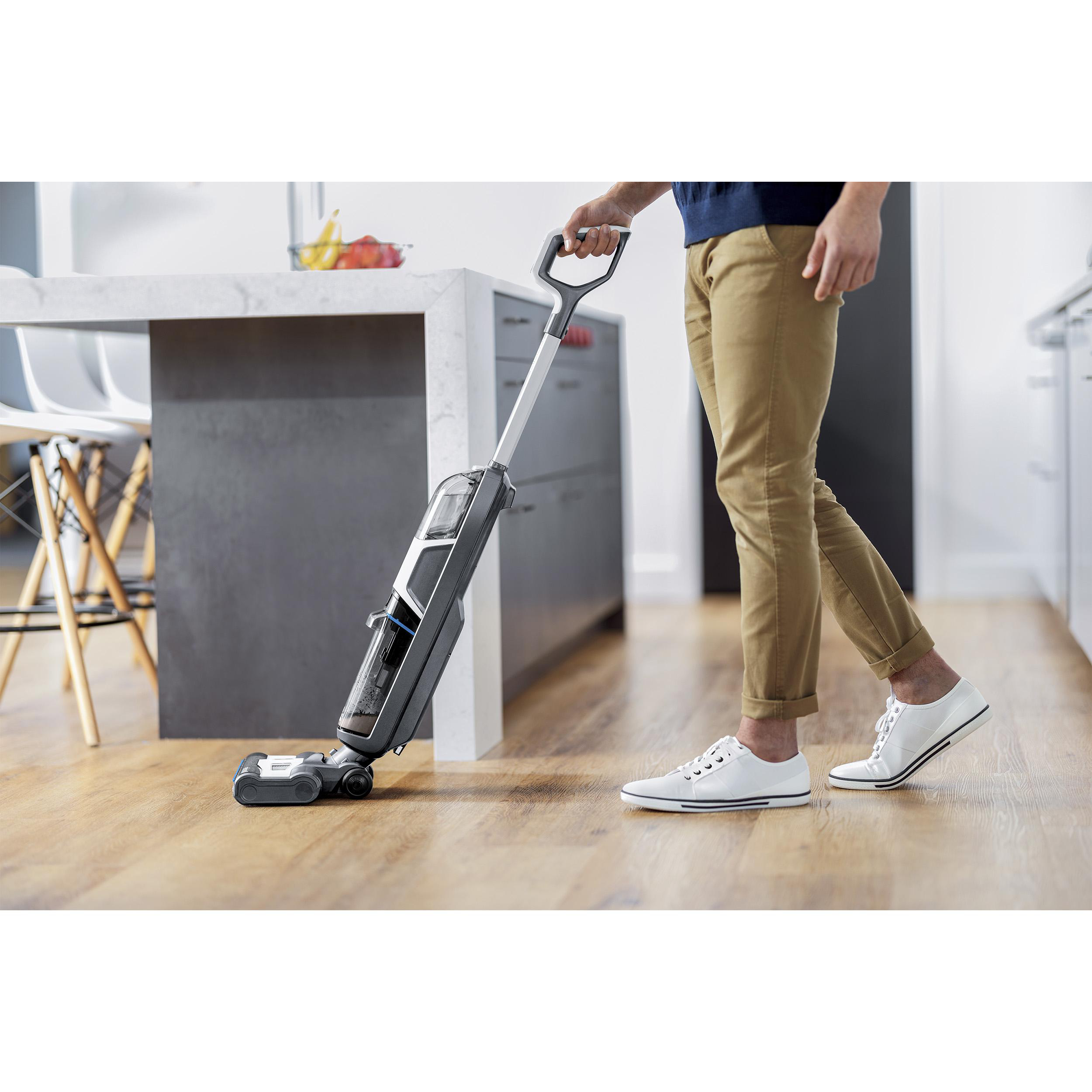 Eine Person saugt einen Holzboden mit einem grau-schwarzen Staubsauger in einer Küche. Weiße Schuhe und Hosen sind sichtbar.
