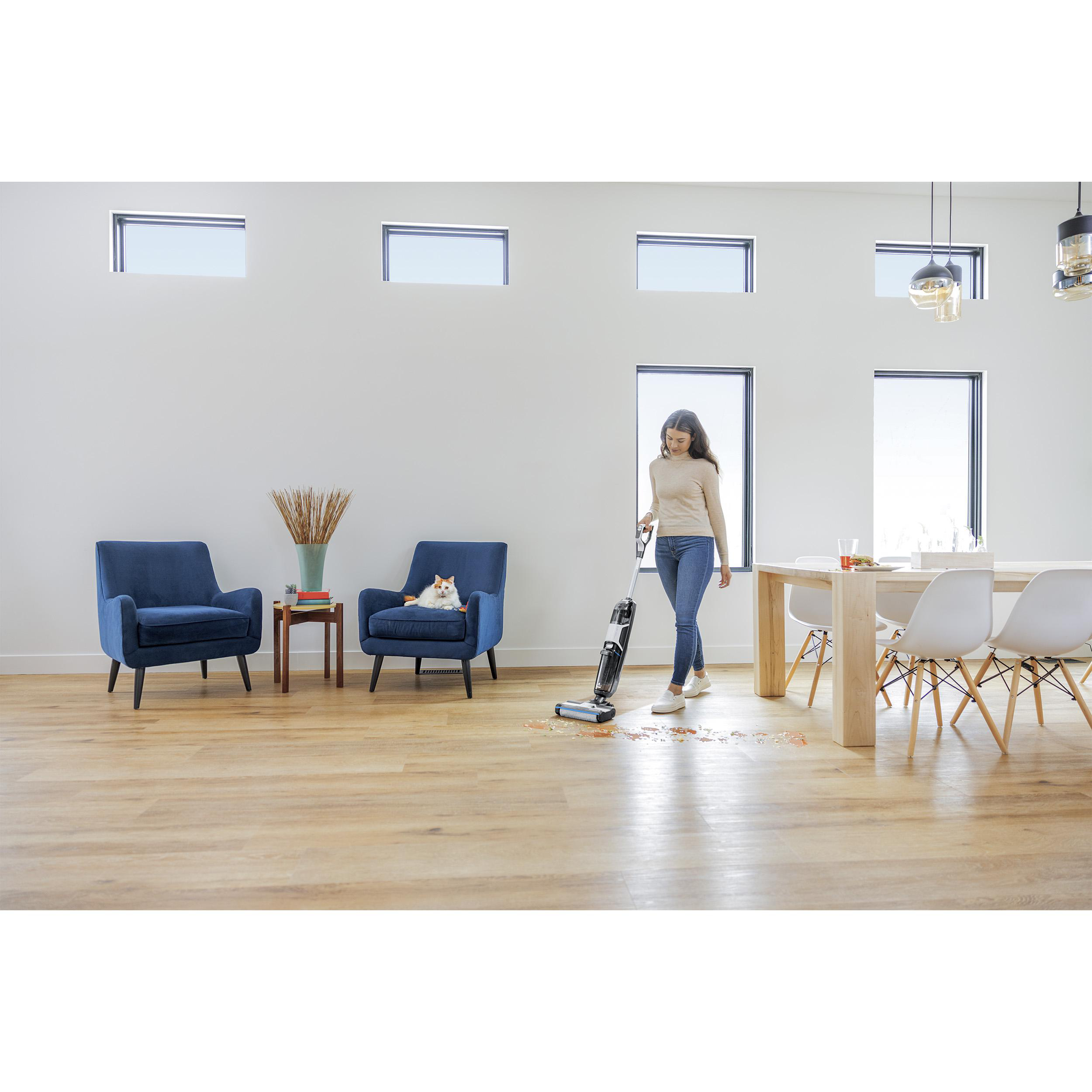Eine Frau saugt einen Holzboden in einem modernen Zuhause. Eine Katze sitzt auf einem blauen Stuhl, und ein Tisch ist gedeckt.