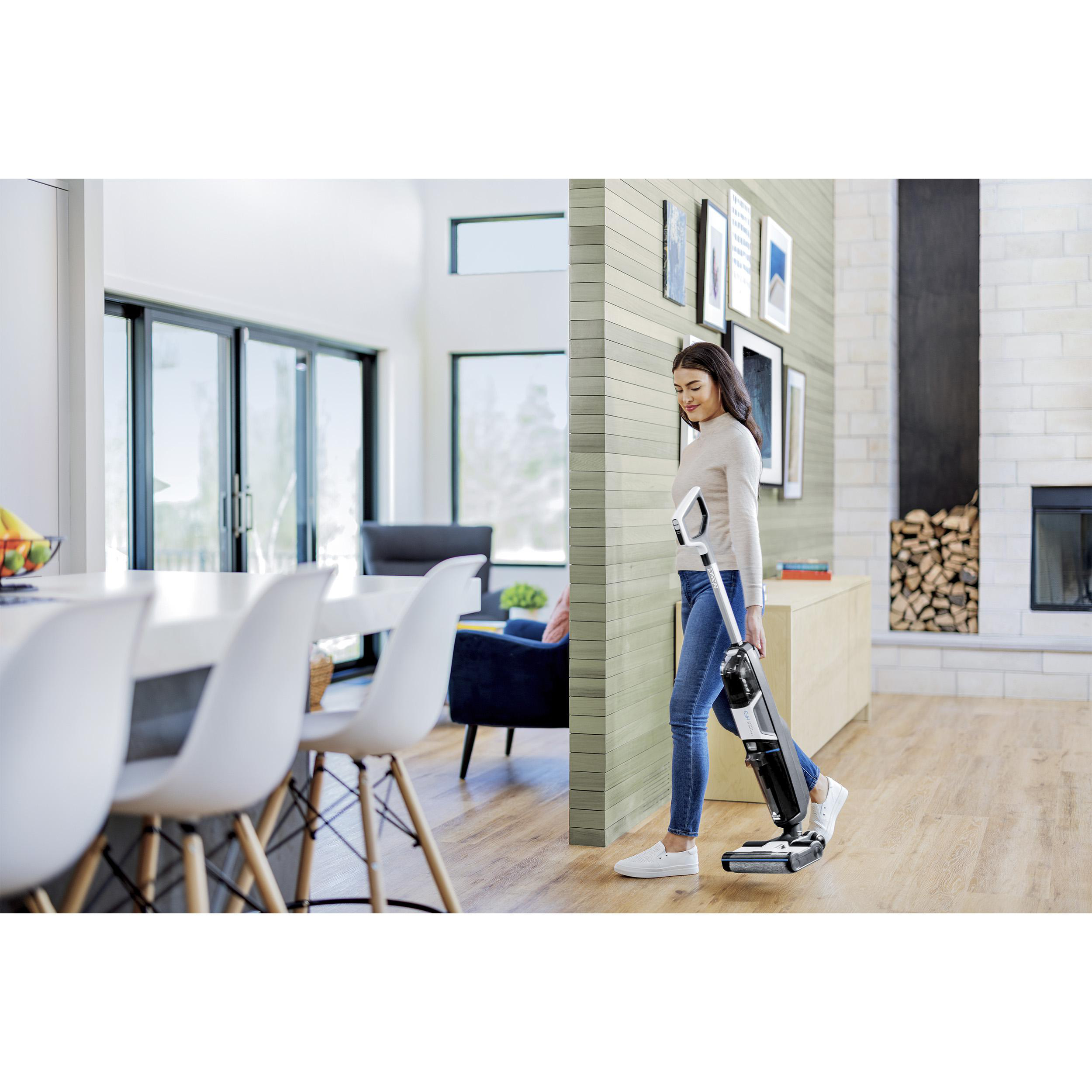 Frau saugt Holzboden in modernem Zuhause. Weiße Stühle, Tisch und blauer Sessel sind sichtbar.