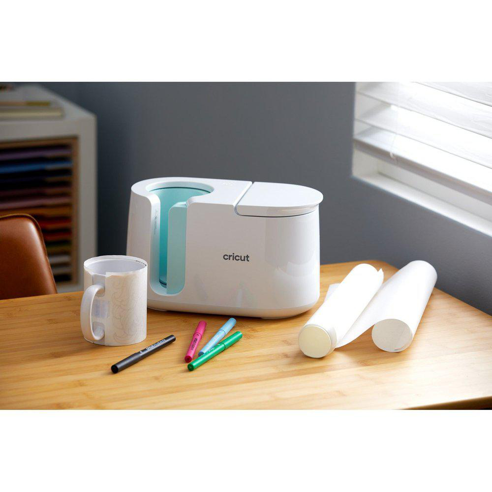 Eine weiße Cricut-Tassenpresse, eine Tasse, Stifte und Papierrollen stehen auf einem Holztisch. Der Hintergrund zeigt ein Fenster und Regale.