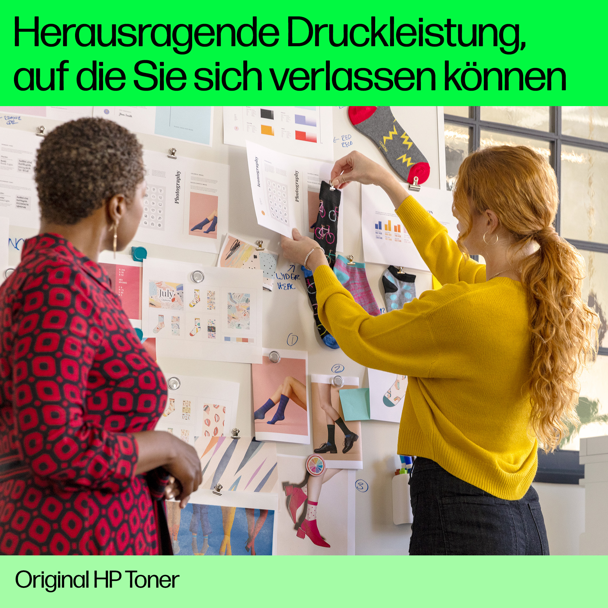 Zwei Frauen in einem Raum betrachten eine Tafel mit Socken und Fotos. Der Hintergrund ist eine Wand.