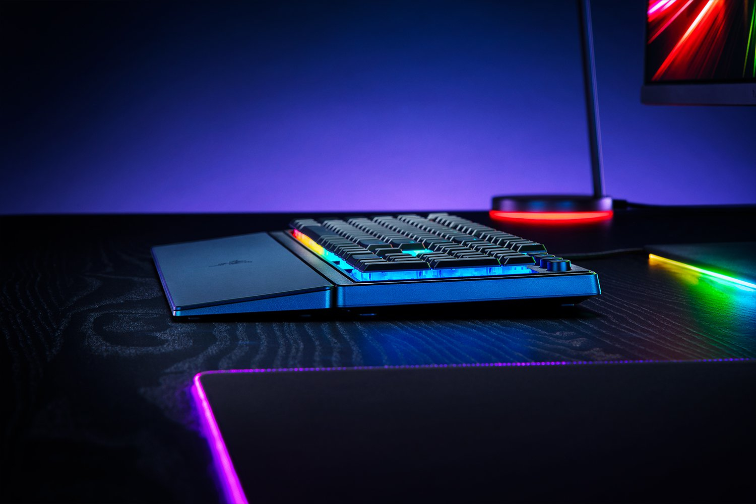 A black gaming keyboard with colorful lights. The keyboard is on a black desk. Purple background.