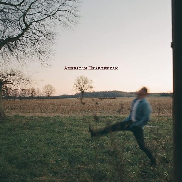 Ein Mann tritt in einem Feld mit dem Text "American Heartbreak". Bewölkter Himmel, Bäume und Feld sind sichtbar.