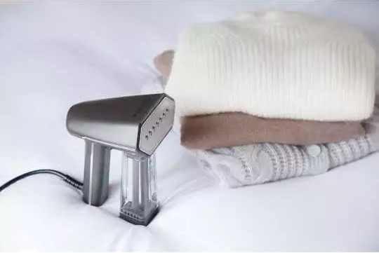 Handheld steamer on a bed with folded sweaters. Steamer is silver and black. Sweaters are white, brown, and gray.