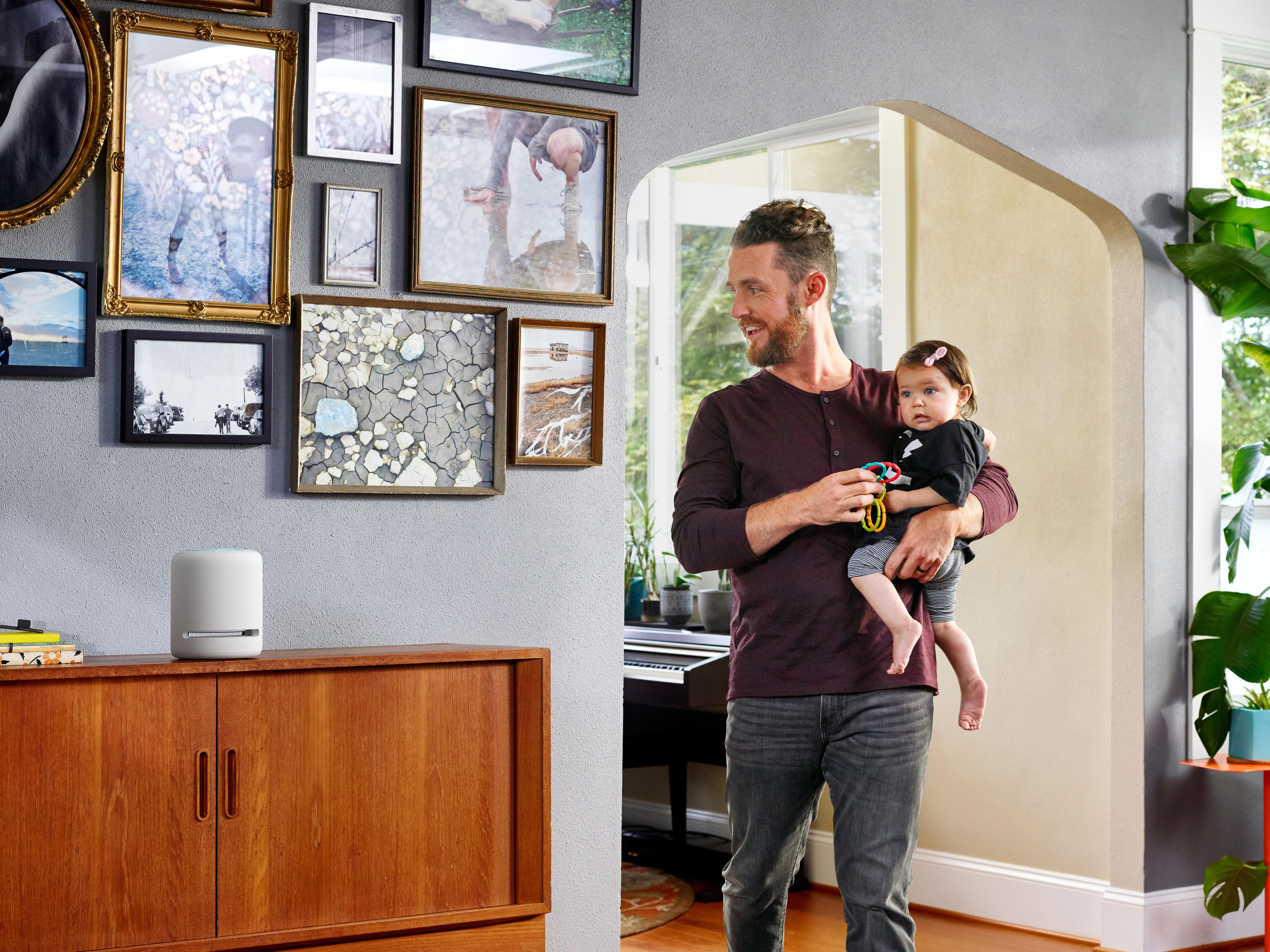 Ein Mann hält ein Baby in einem Raum mit Kunstwerken an der Wand. Ein intelligenter Lautsprecher steht auf einem Holzschrank.