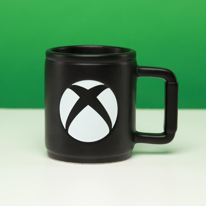Une tasse noire avec le logo Xbox en blanc. La tasse est sur une surface blanche, fond vert.