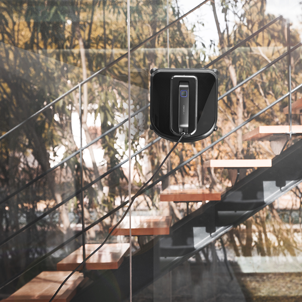 Un robot limpiador de ventanas negro en una ventana de vidrio con una escalera visible detrás.