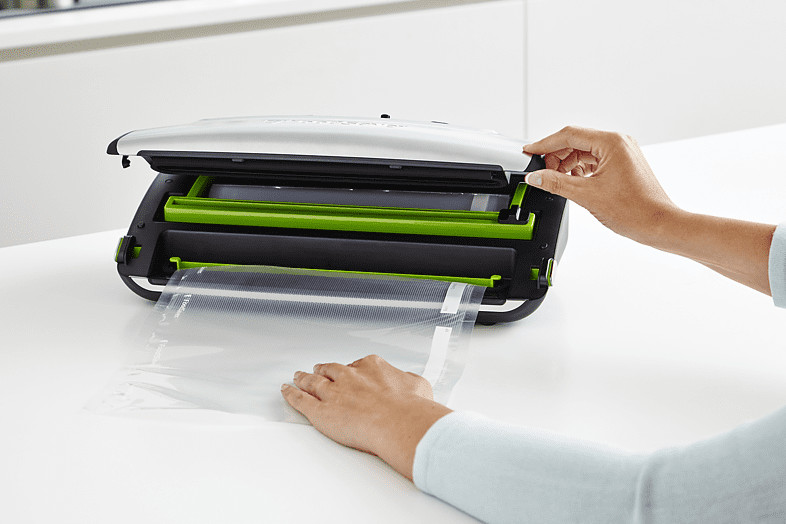 Persoon die een voedselverzegelaar gebruikt. Machine is zwart en groen. Transparante plastic zak wordt ingebracht.
