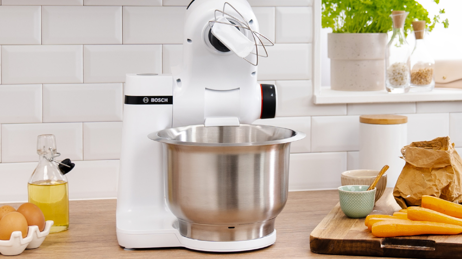 Weißer Küchenmixer mit Schüssel und Aufsätzen auf der Küchentheke. Zutaten sichtbar.