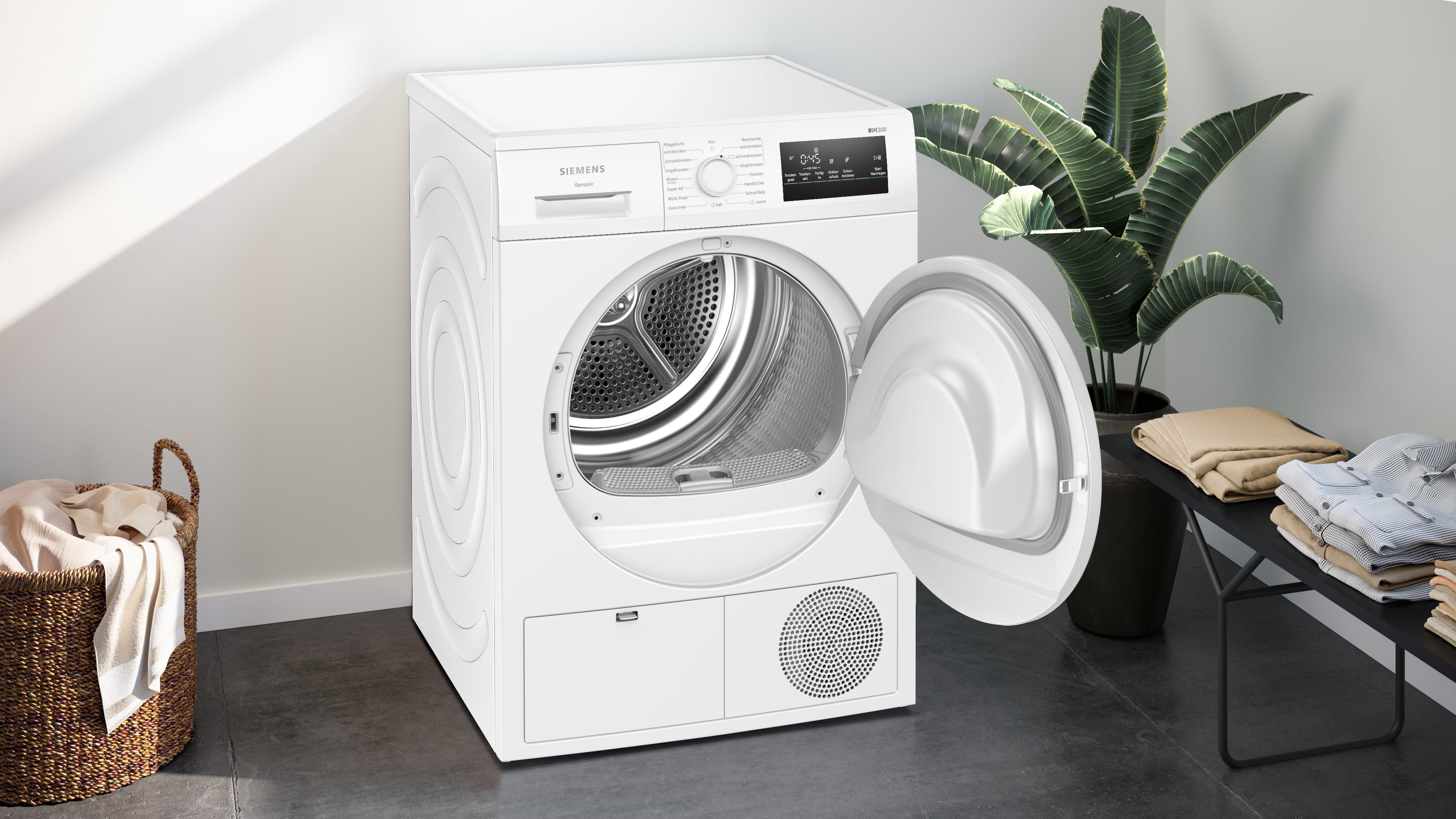 Ein weißer Siemens-Trockner mit offener Tür. Eine Pflanze, Kleidung und ein Korb befinden sich in der Nähe.