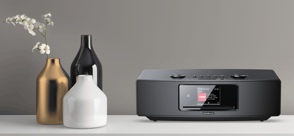 Ein schwarzes Kenwood Internetradio mit Display, in der Nähe von Vasen und Blumen.