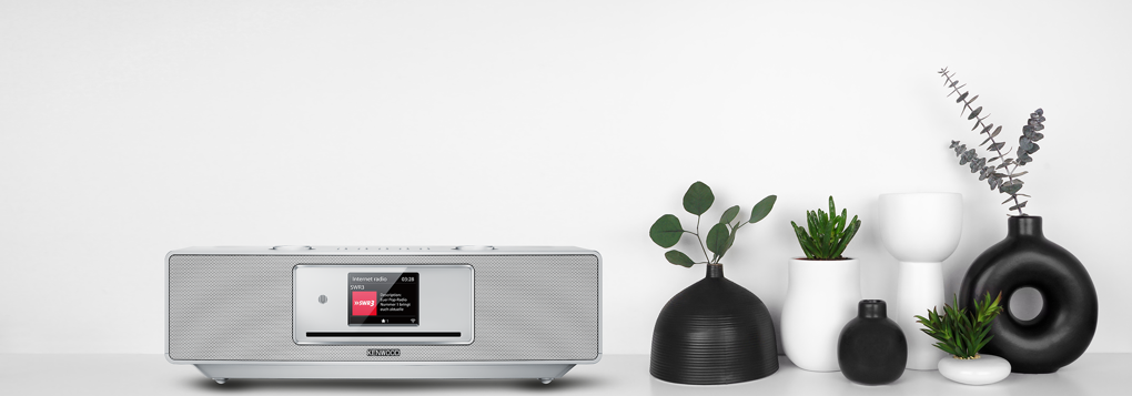 Ein silbernes Radio mit einem Bildschirm und eine Reihe von Vasen mit Pflanzen. Der Hintergrund ist weiß.