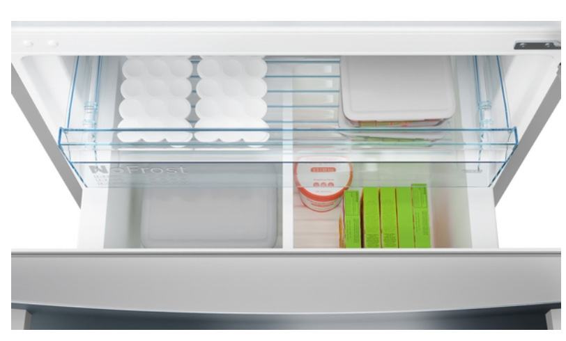 Cajón de refrigerador abierto con bandejas de hielo, recipientes de alimentos y artículos empaquetados. El interior es blanco y organizado.