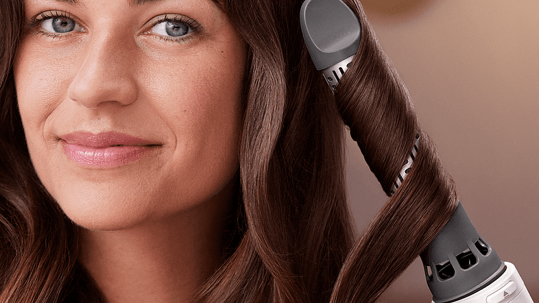 Woman curling her hair with a curler. She is looking at the camera and smiling.