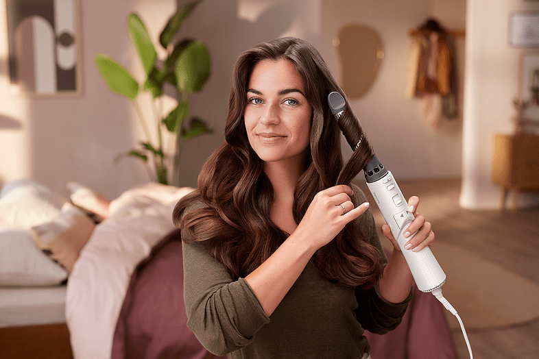 Woman styling her hair with a curling iron, smiling at the camera.