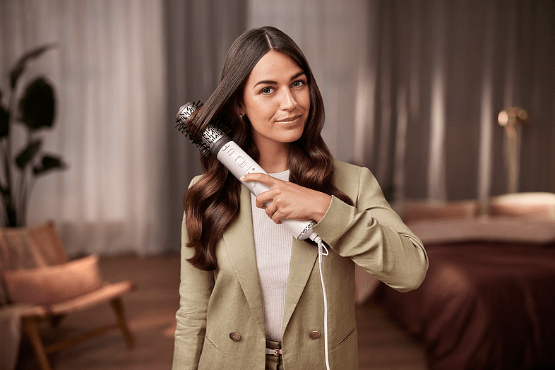 A woman with long brown hair is styling her hair with a styling brush.