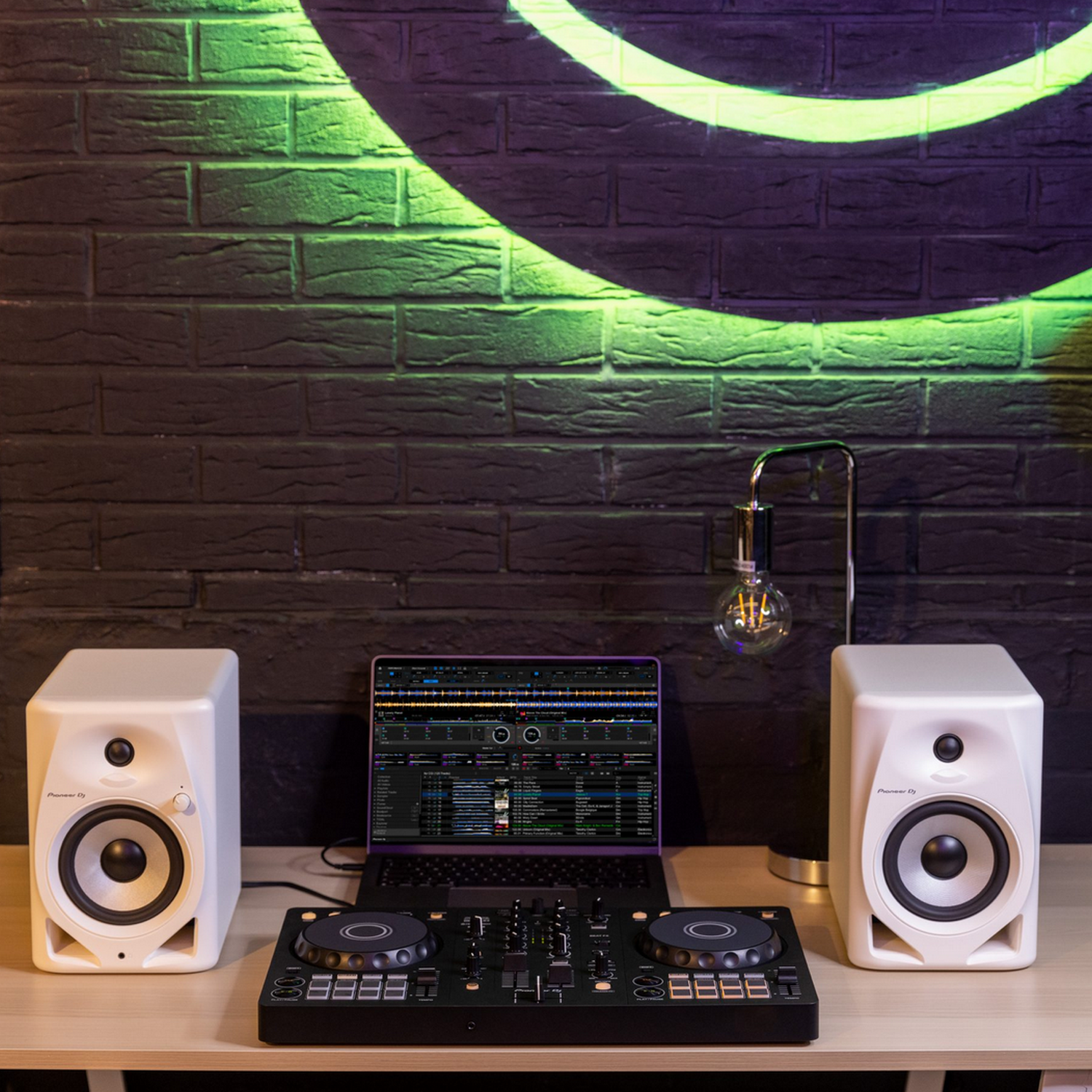 DJ-opstelling met luidsprekers, een laptop en een mixer op een houten tafel, tegen een zwarte bakstenen muur.