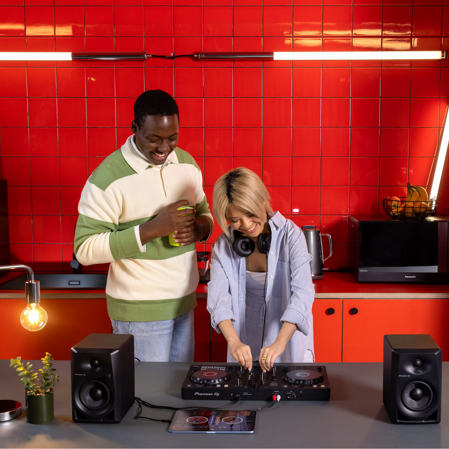Twee mensen in een keuken. Eén bedient een DJ-mixer. Rode tegel achtergrond.
