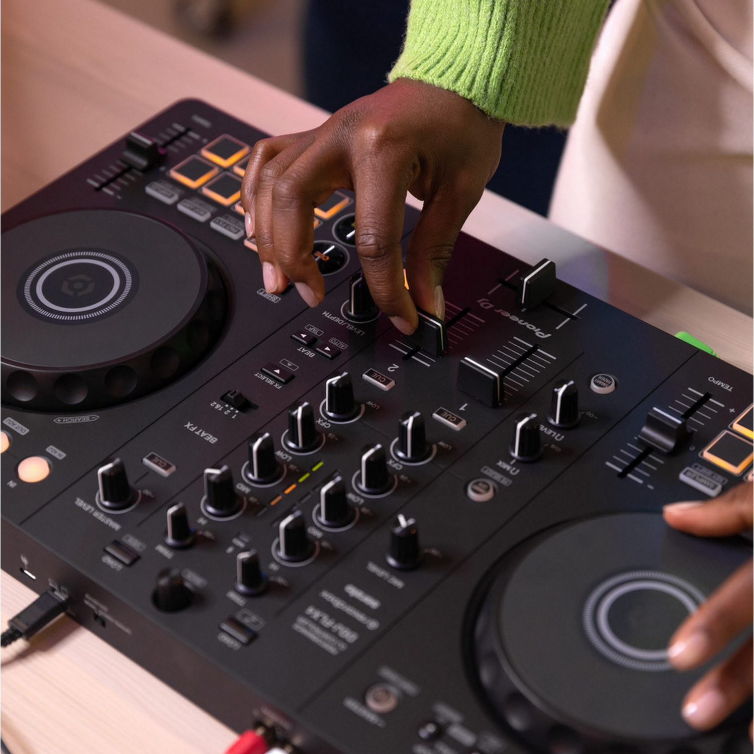 Handen van een persoon die een zwarte DJ-mixer aanpast. De persoon draagt een groene trui.