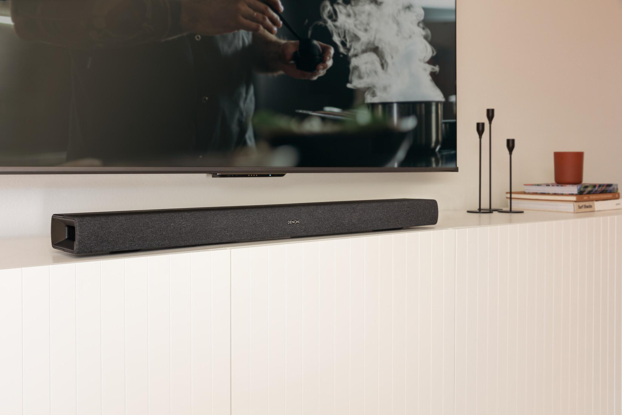 Eine schwarze Soundbar unter einem Fernseher auf einem weißen Schrank. Kerzen und Bücher sind rechts.