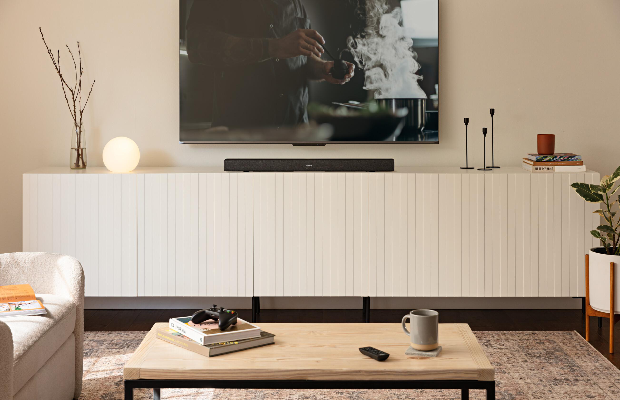 Wohnzimmer mit Fernseher, Soundbar, weißem Schrank, Couchtisch mit Deko. Couch links.