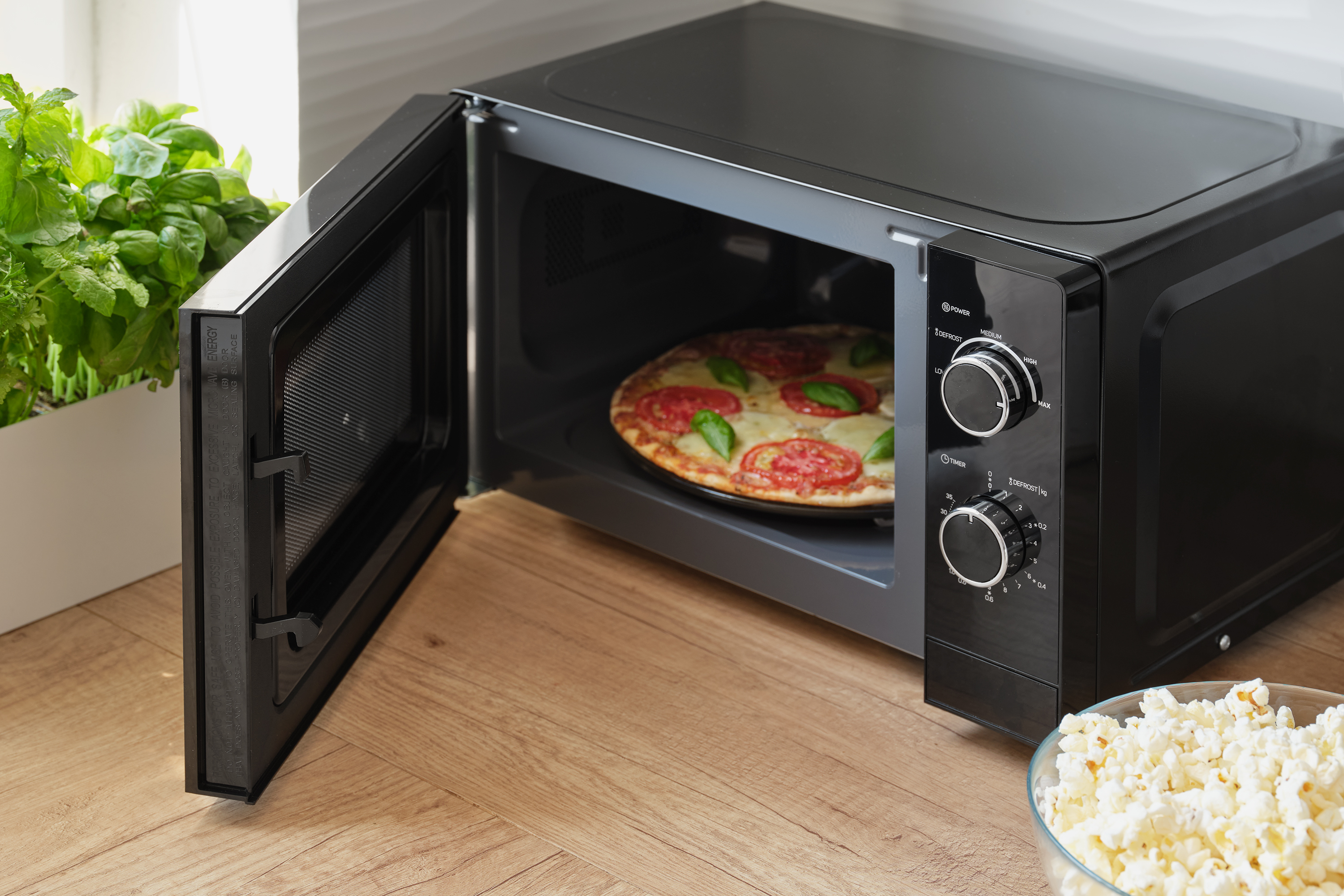 Fekete mikrohullámú sütő nyitott ajtóval, benne pizzával. Popcorn egy tálban a közelben.
