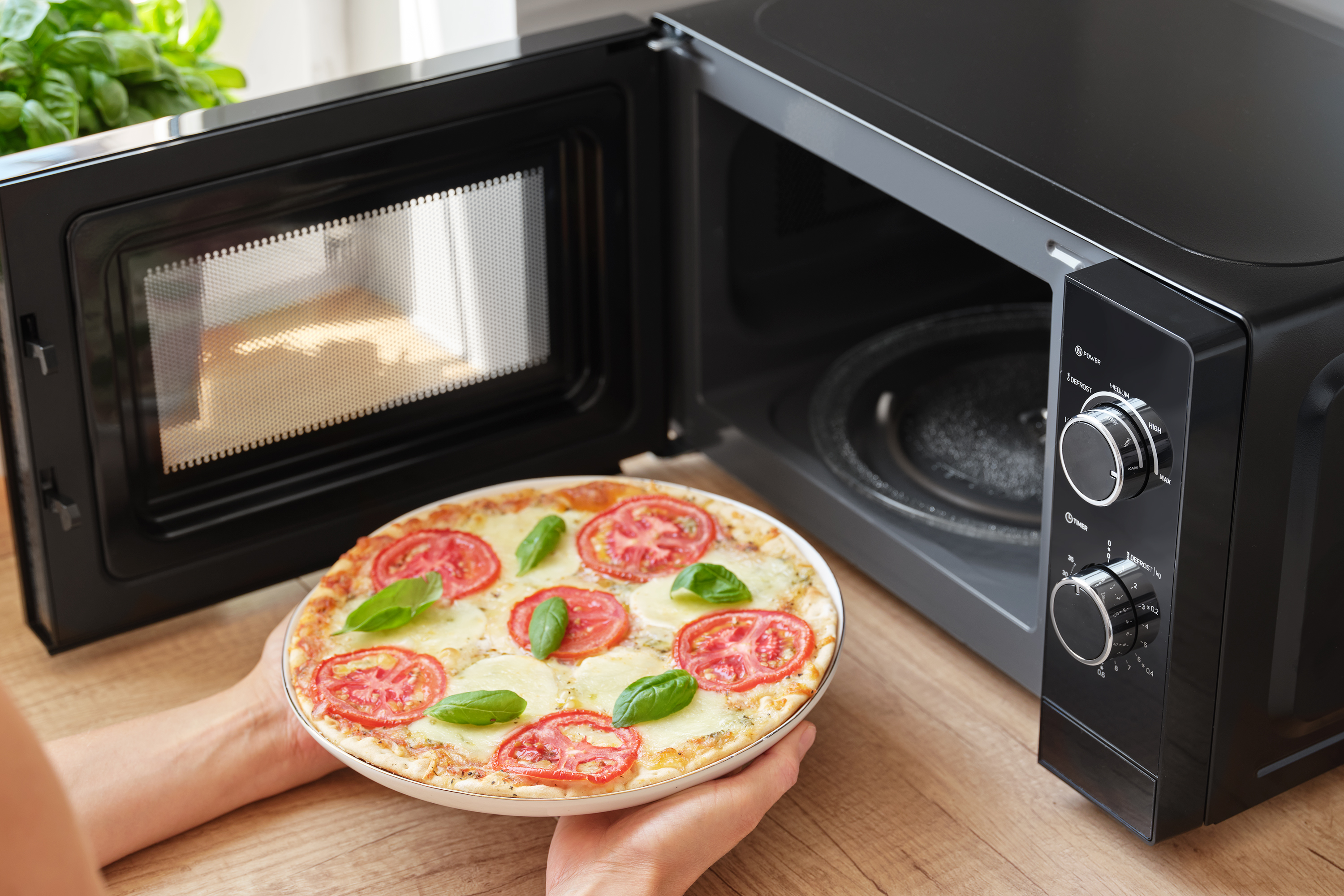 Pizza, amelyet kezek tartanak egy nyitott fekete mikrohullámú sütő mellett.