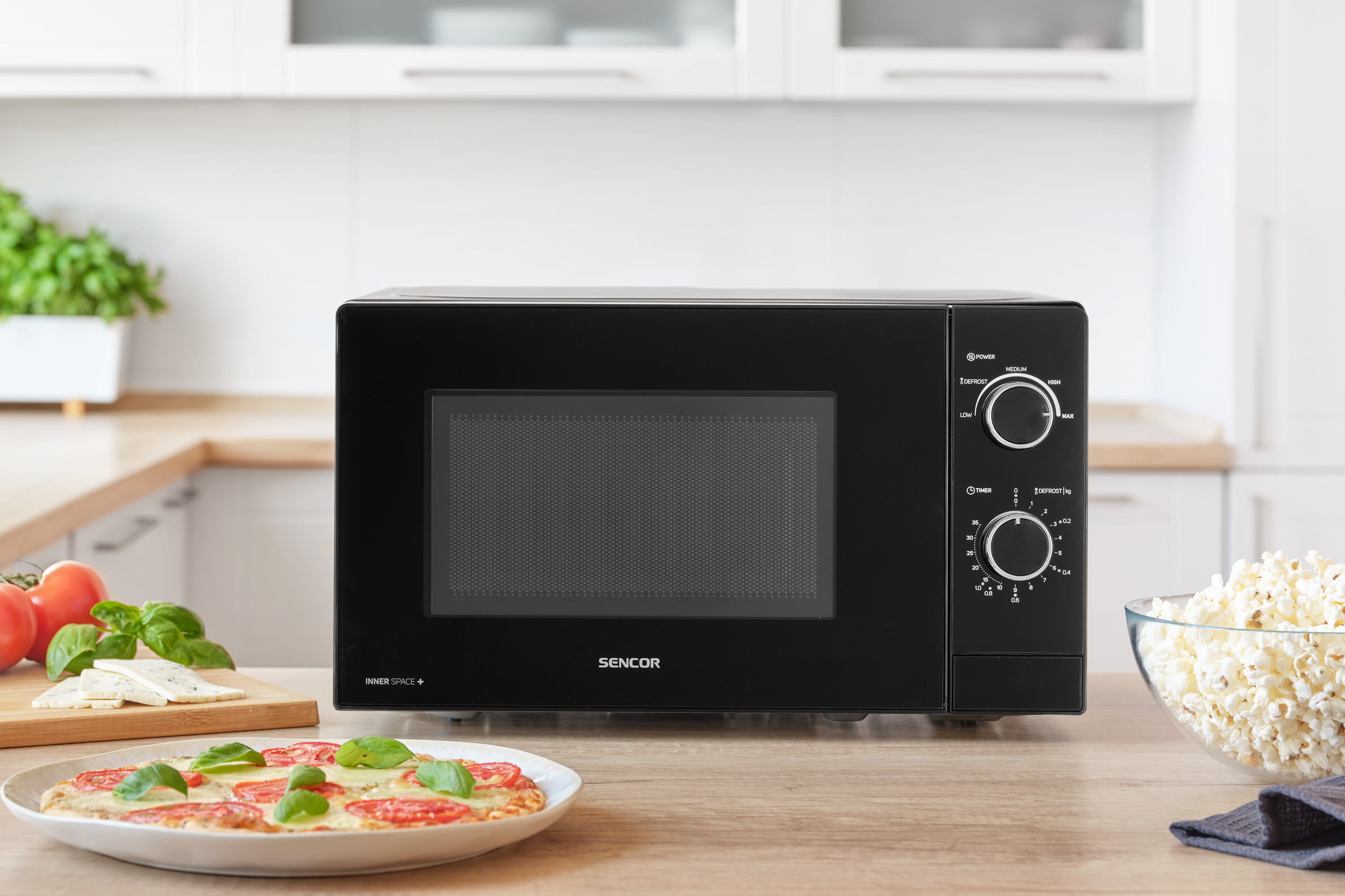 Fekete mikrohullámú sütő fa felületen, pizza, sajt, pattogatott kukorica, vágódeszka a közelben.