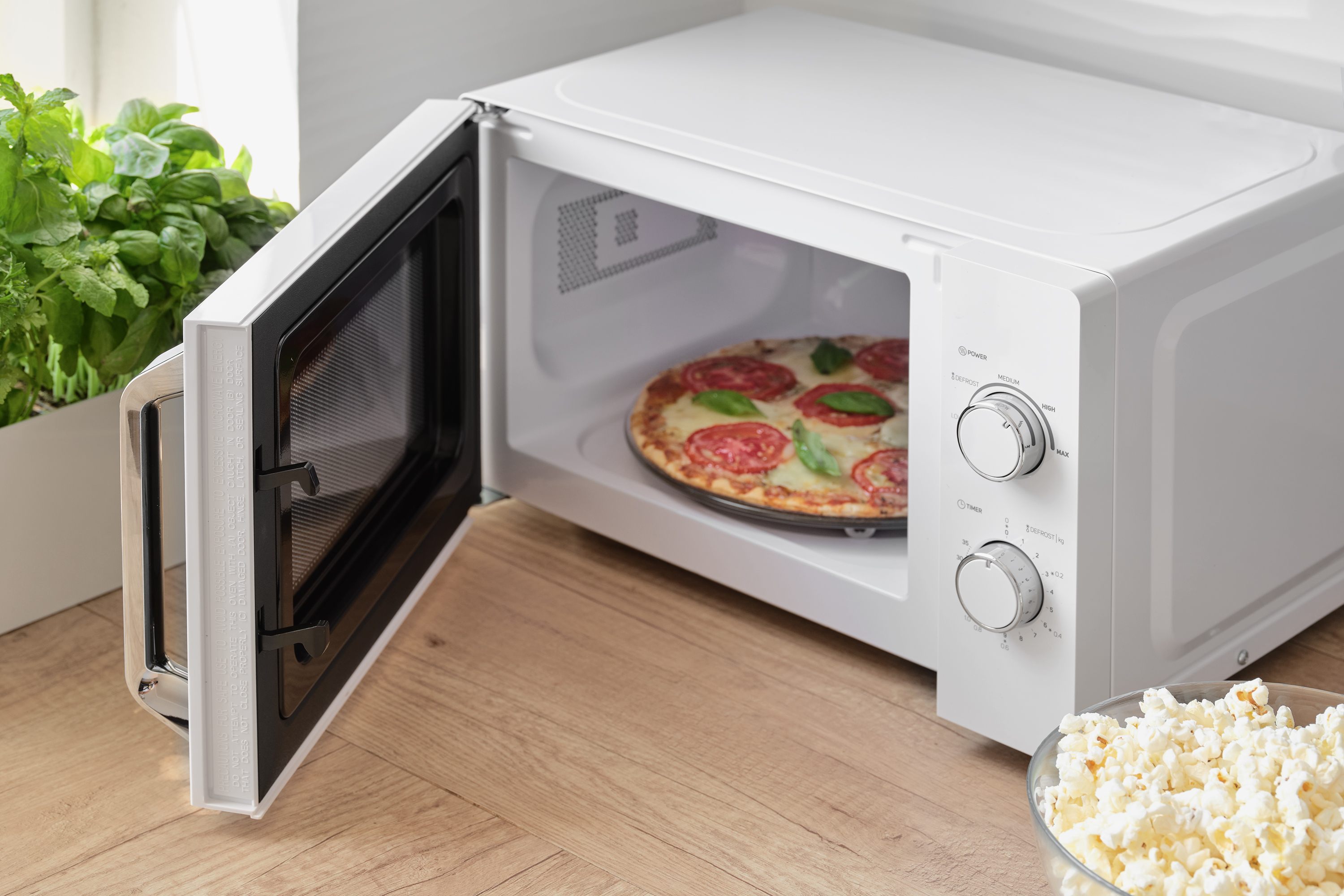 Fehér mikrohullámú sütő nyitott ajtóval, pizzát mutat. A jobb oldalon egy tál pattogatott kukorica.