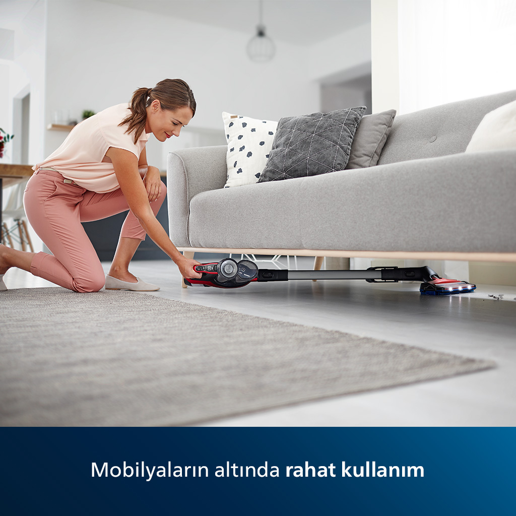 Gri bir halı üzerinde gri bir kanepenin altında kırmızı ve siyah kablosuz bir elektrikli süpürge kullanan kadın.