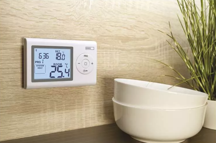 Egy falra szerelt termosztát digitális kijelzővel. Tálak és egy növény a háttérben.