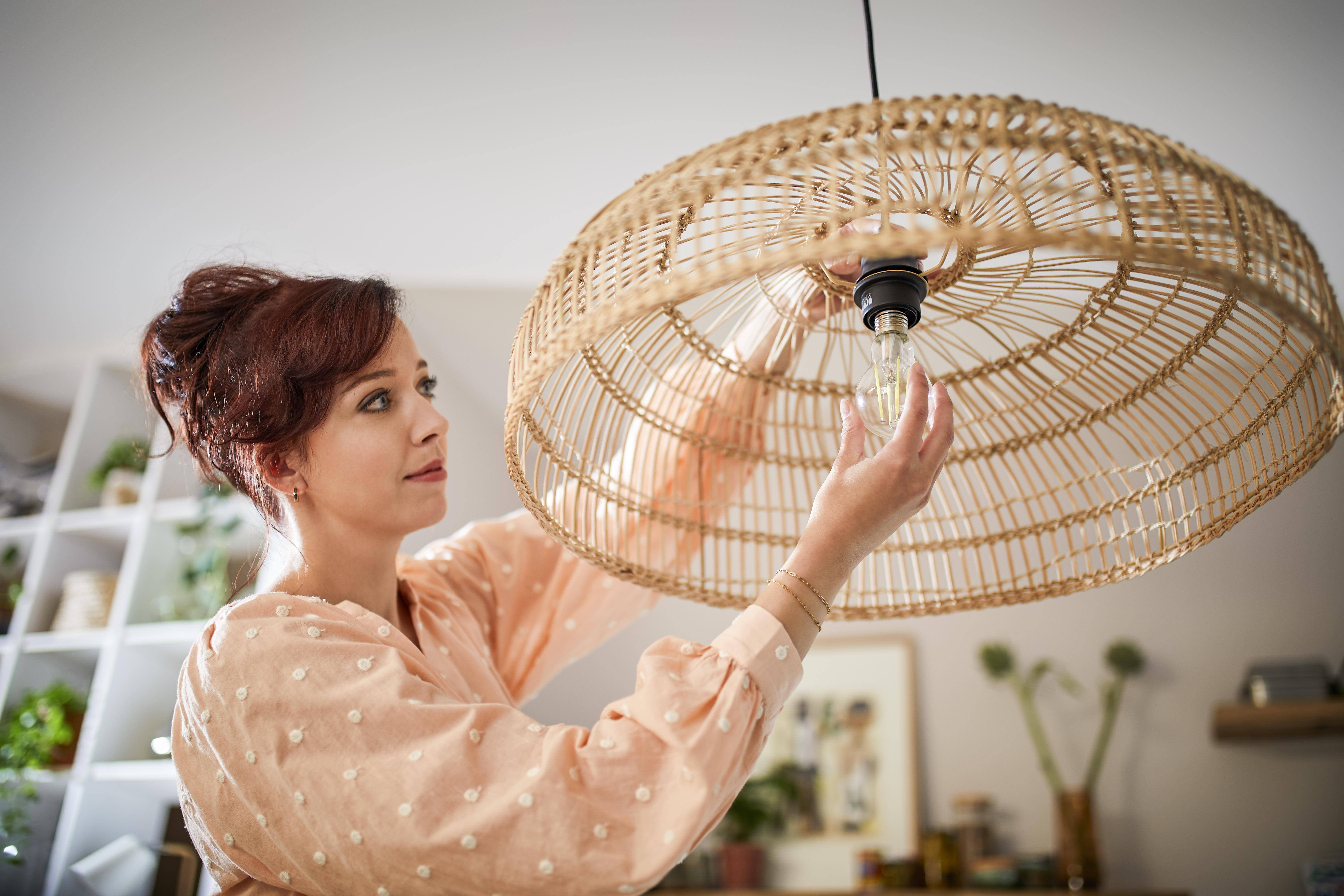 Frau in pfirsichfarbenem Oberteil ersetzt Glühbirne in einem Korblampenschirm. Weiße Regale und Dekoration im Hintergrund.