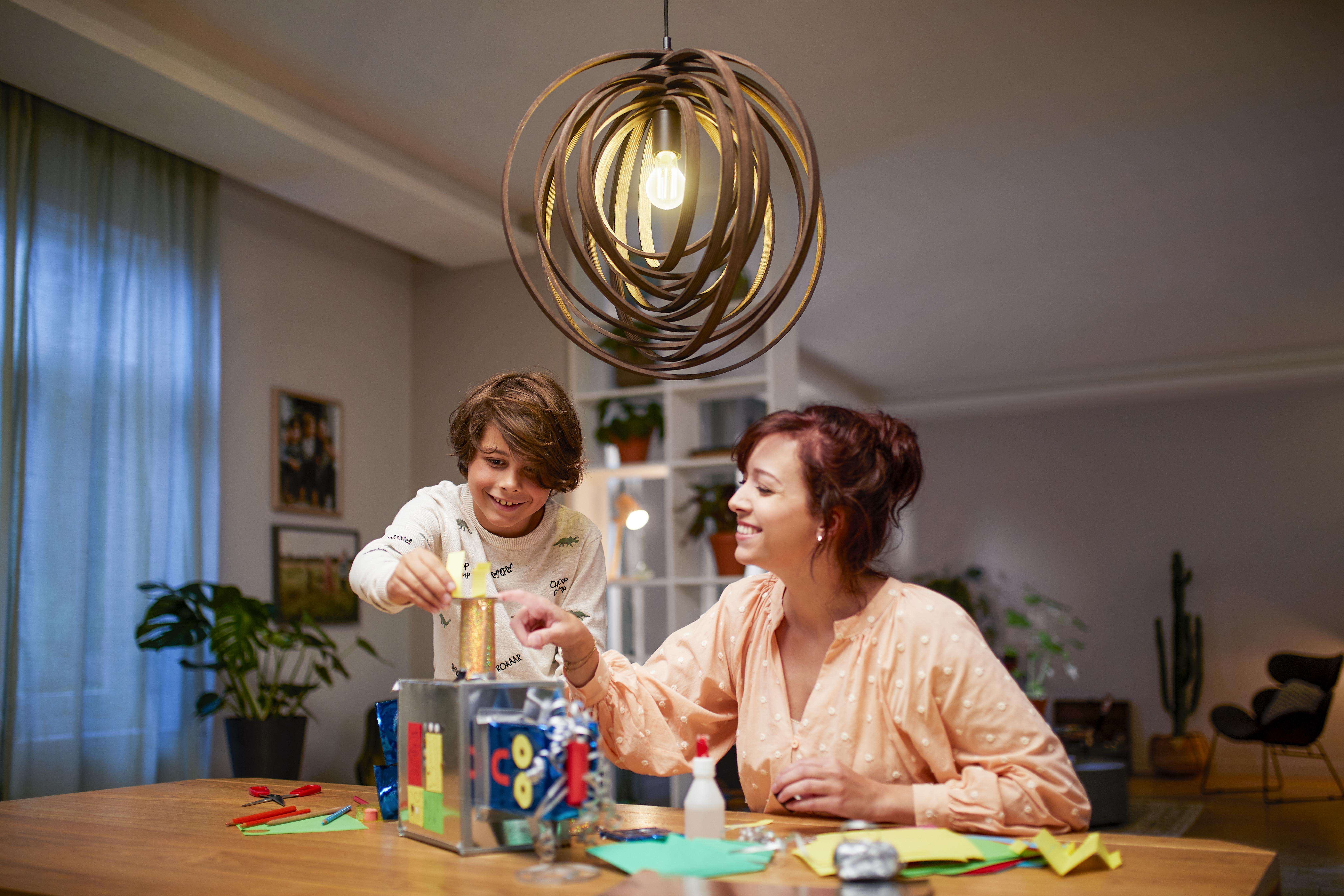 Ein Junge und eine Frau bauen ein Modell unter einer dekorativen Hängeleuchte, drinnen, Holztisch.