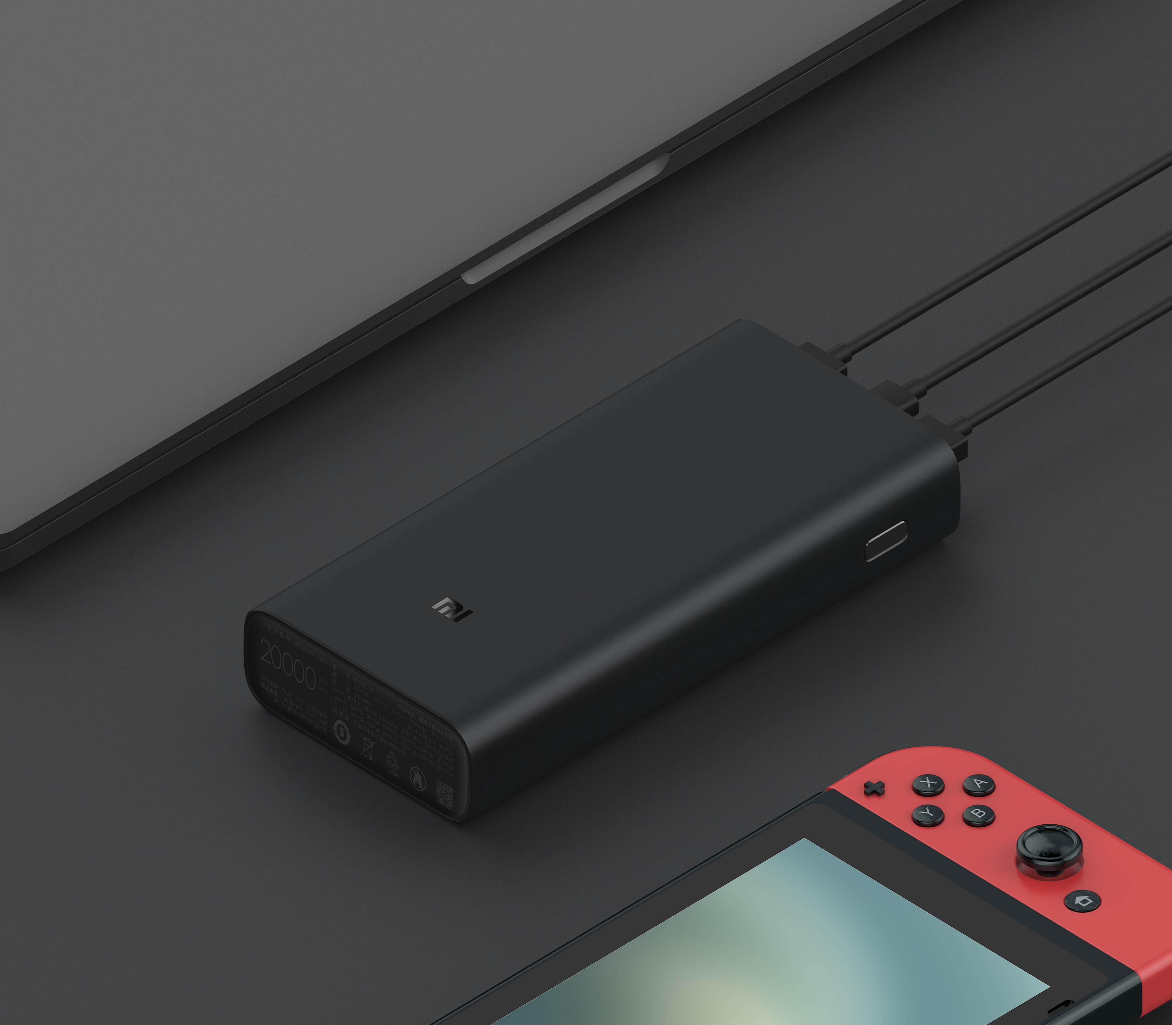 Power bank nero che carica dispositivi. Laptop e console su superficie scura.