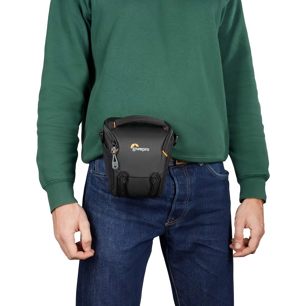 Eine Person trägt einen grünen Pullover und blaue Jeans mit einem schwarzen Lowepro-Kameraholster am Gürtel.