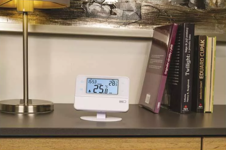 Fehér digitális termosztát asztalon, könyvek, lámpa, sötét háttér.