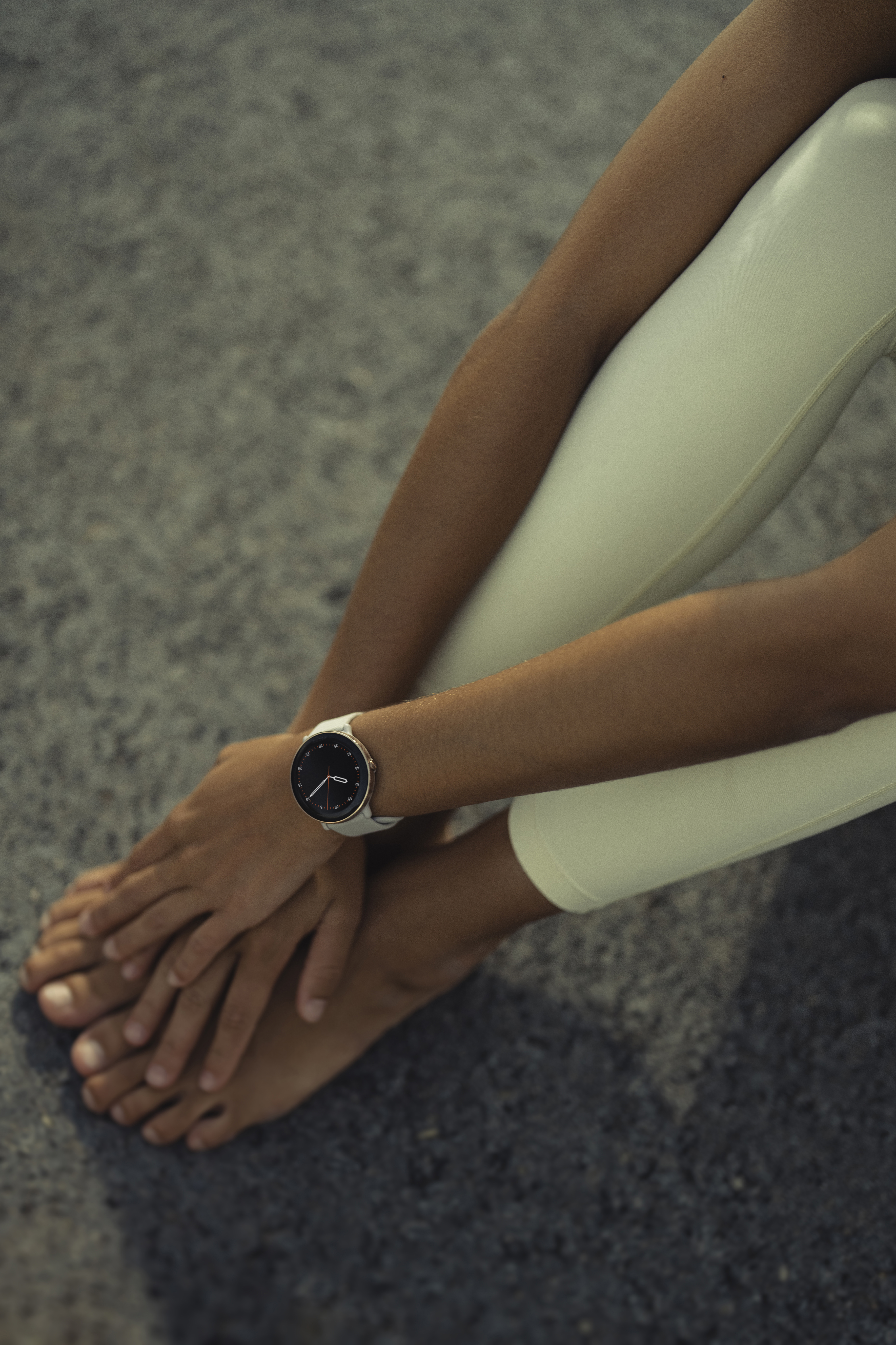 Mujer con reloj inteligente en la muñeca. Sentada, piernas y pies visibles, sobre superficie gris.