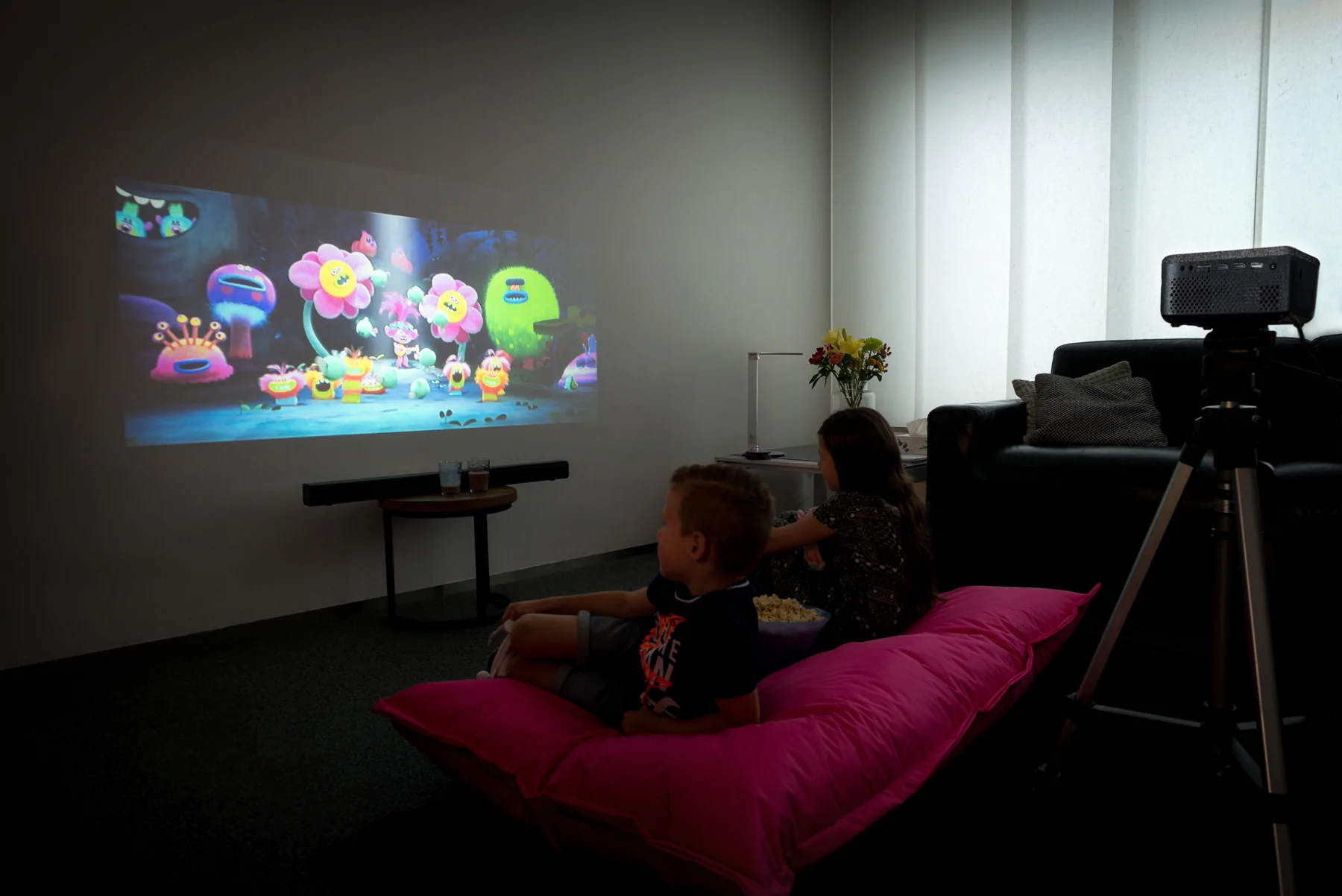 Deux enfants regardent un film sur un mur blanc. Un projecteur diffuse le film. Un pouf rose est au premier plan.