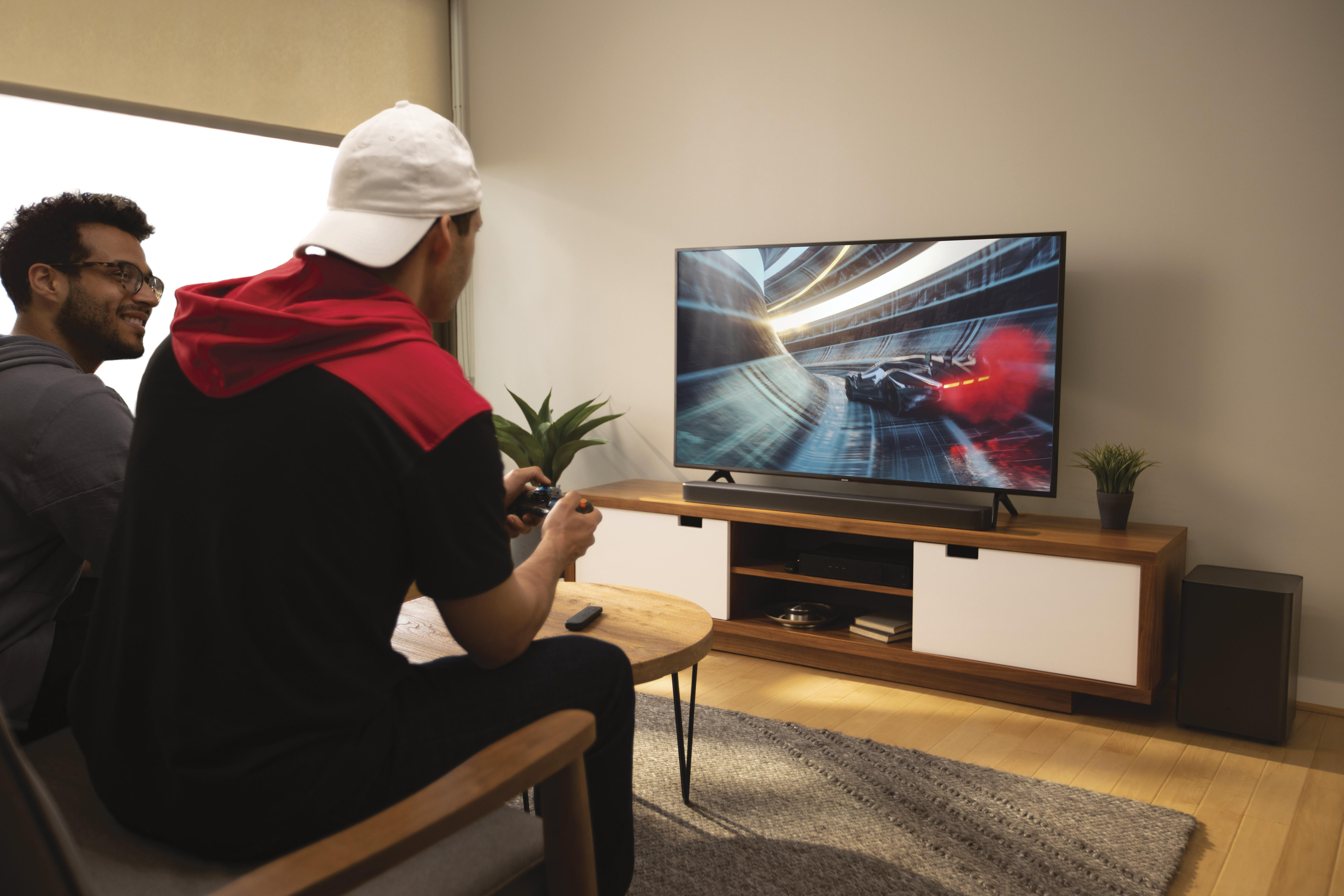 Zwei Männer spielen im Wohnzimmer. TV zeigt Rennspiel. Soundbar auf dem Schrank darunter. Subwoofer.
