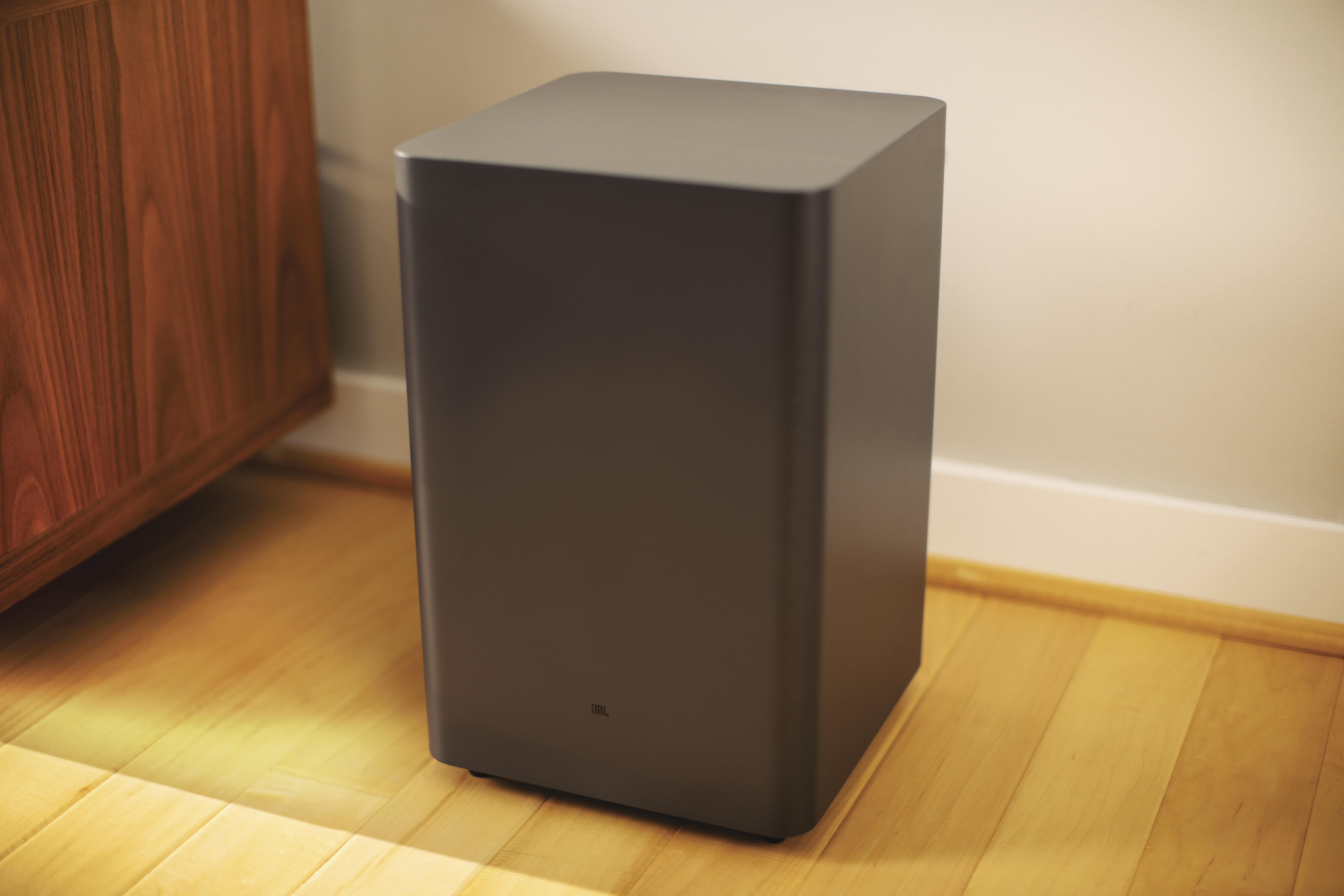 Ein dunkelgrauer Subwoofer auf hellem Holzboden, neben einem Holzschrank und einer weißen Wand.
